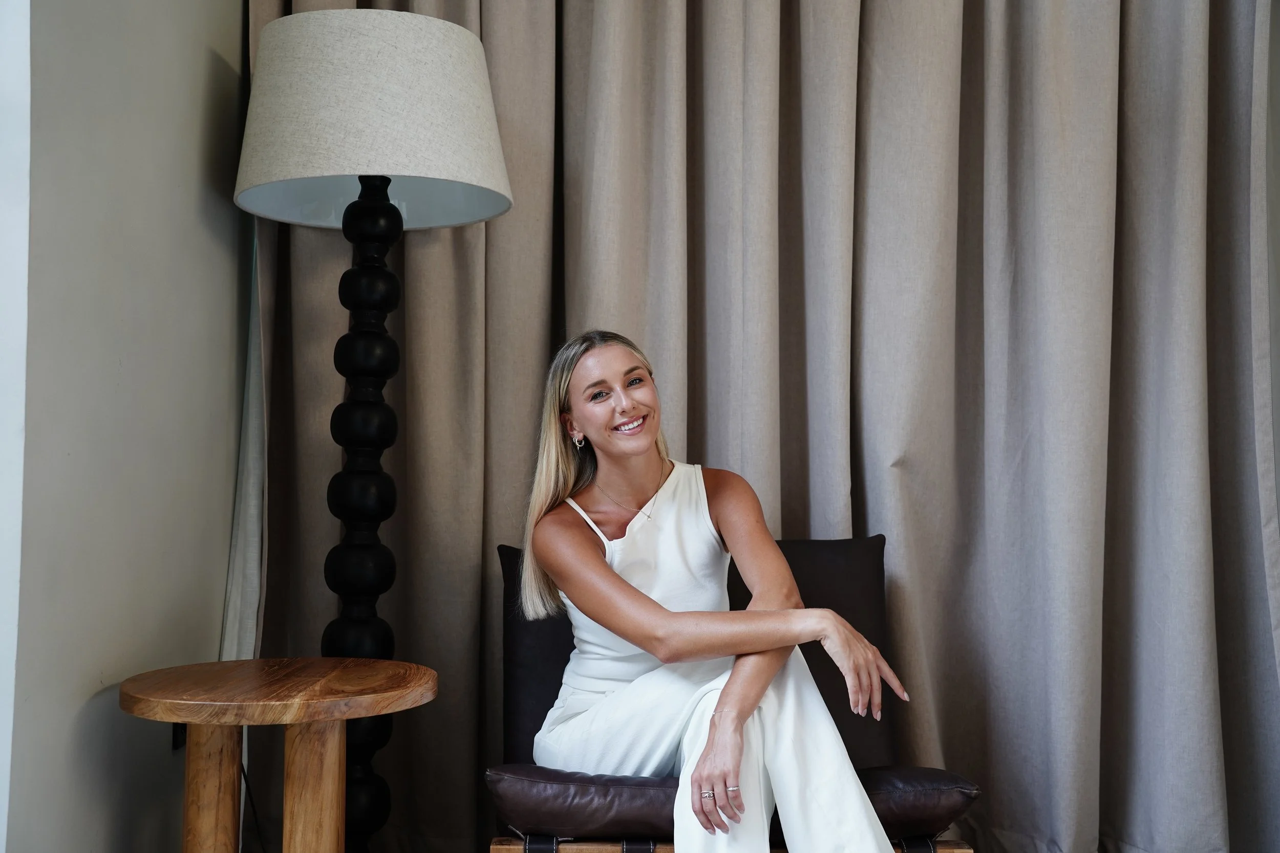 A woman with long blonde hair sitting on a chair in front of beige curtains, smiling at the camera, wearing a white sleeveless dress.
