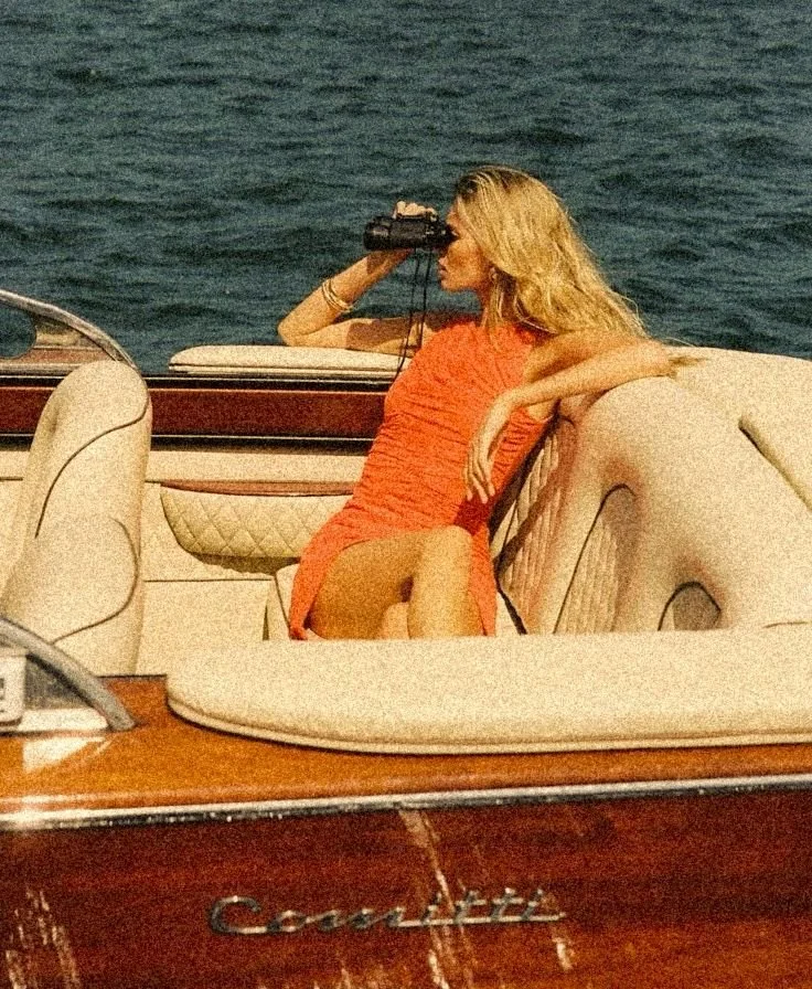 A woman with blonde hair wearing an orange dress sitting on a boat, looking through binoculars towards the water.