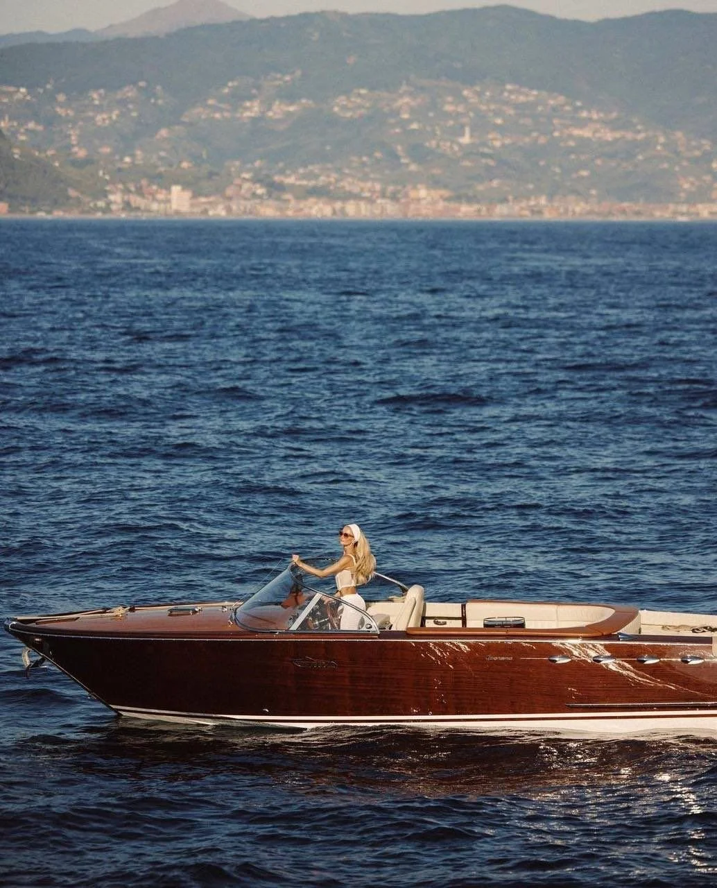 A woman with long blonde hair and sunglasses sits on a luxury boat with a polished wooden exterior, cruising on a large body of water with a distant shoreline and mountains in the background.