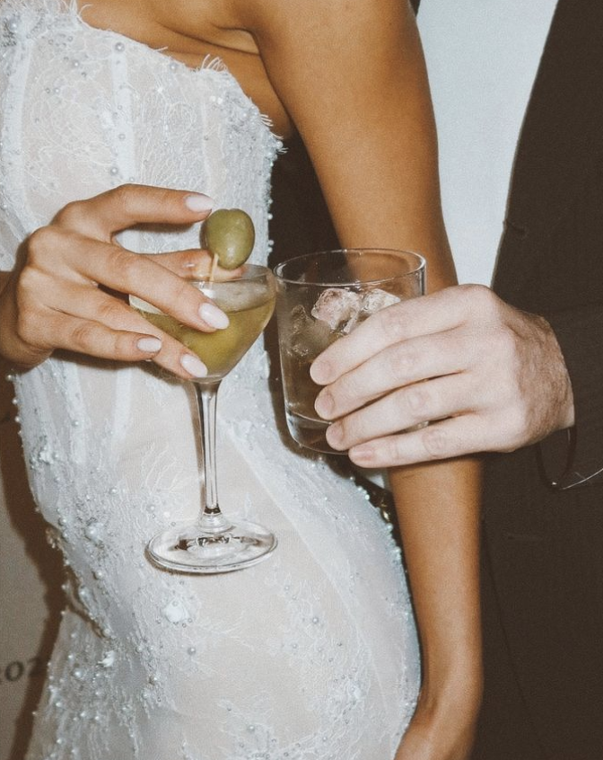 Close-up of a bride and groom holding glasses of drinks at a wedding celebration. The bride has white lace wedding dress and is holding a glass of green olive cocktail. The groom is wearing a dark suit and holding a glass of whiskey with ice.