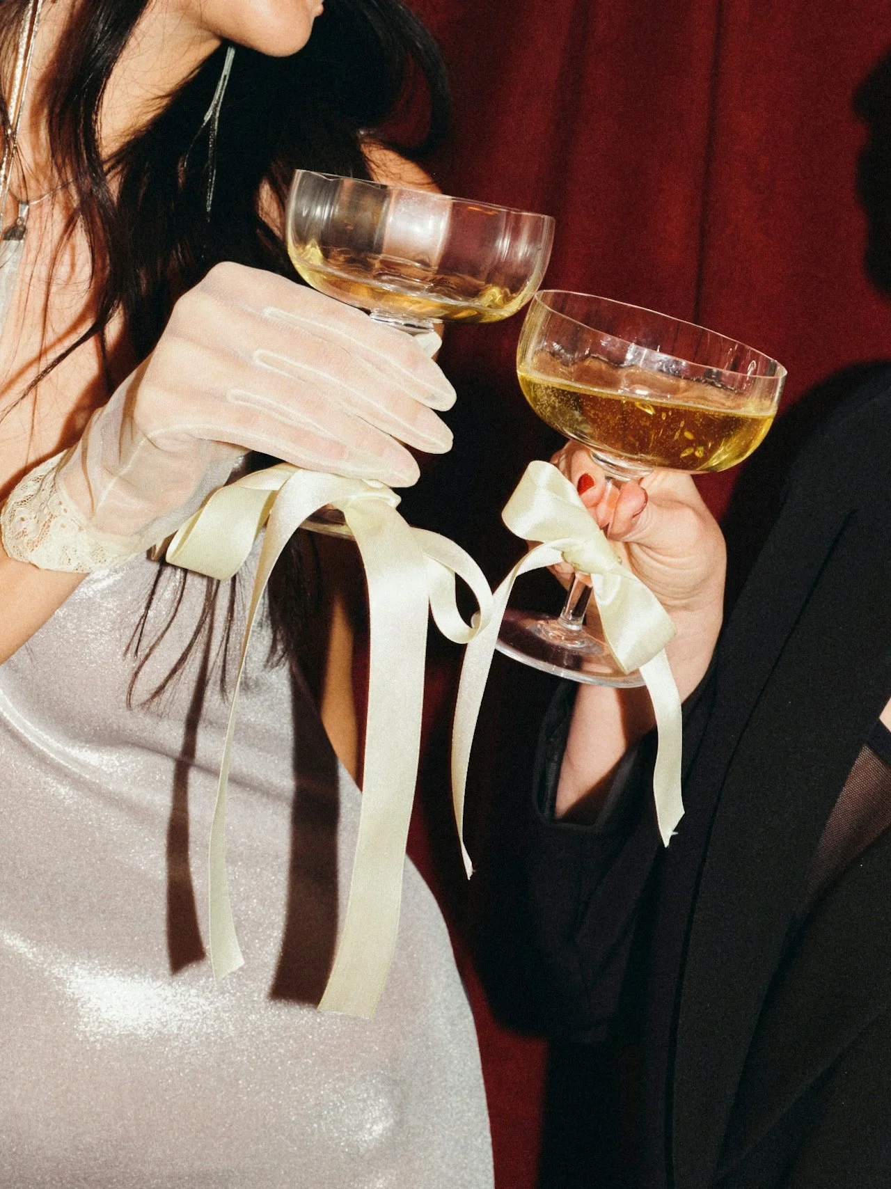 Two people celebrating with glasses of champagne. One person is wearing a white dress and gloves with ribbon details, and the other is wearing a black suit. The background is dark red.