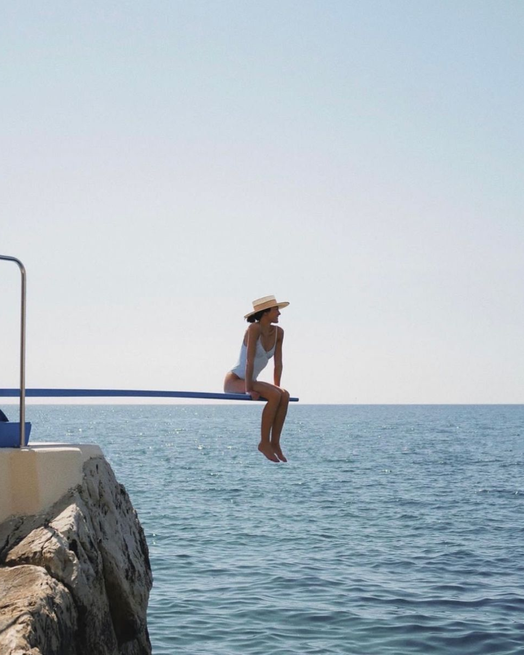 A woman sitting on a diving board over the ocean, wearing a swimsuit and a wide-brimmed hat.