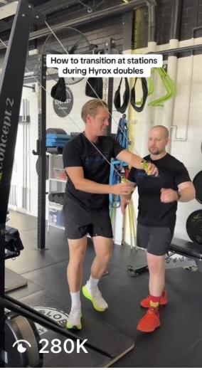 Two men training in a gym with various workout equipment, including resistance bands and weights. One man is demonstrating a resistance band exercise while the other observes. Text overlay reads 'How to transition at stations during Hyrox doubles.'