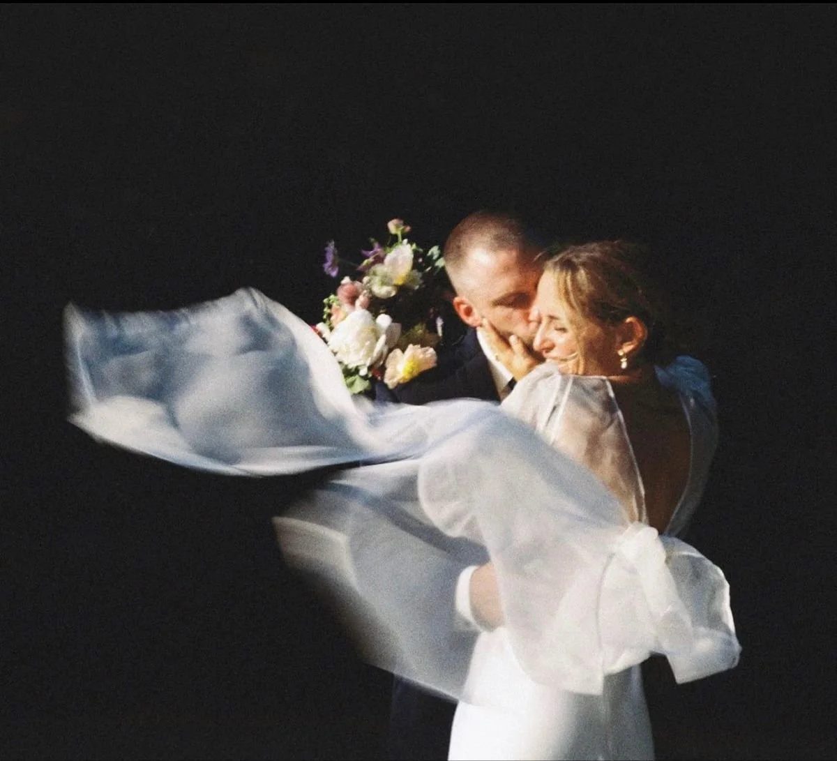 A newlywed couple in wedding attire sharing a romantic moment, with the woman holding a bouquet of flowers, against a dark background.