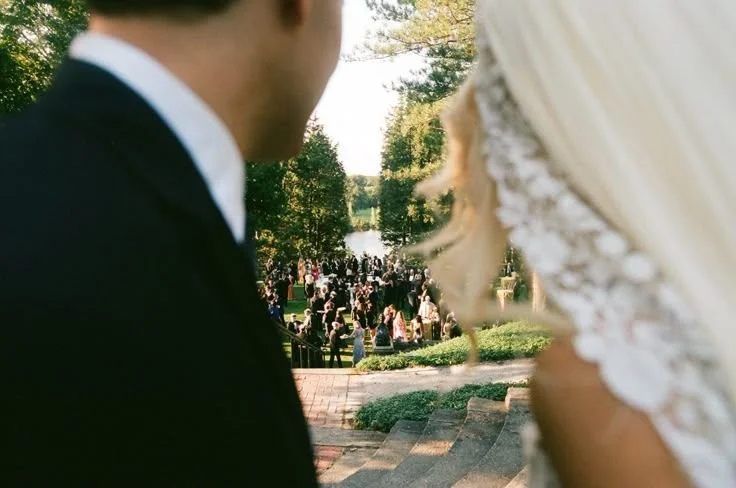 Close-up of a bride and groom at their outdoor wedding ceremony, with guests in the background and trees around.
