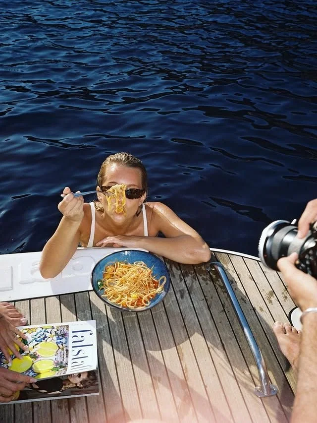 A woman wearing sunglasses is sitting in a boat next to water, holding spaghetti with a fork near her mouth. There is a bowl of spaghetti on the boat and a person holding a camera taking her picture.