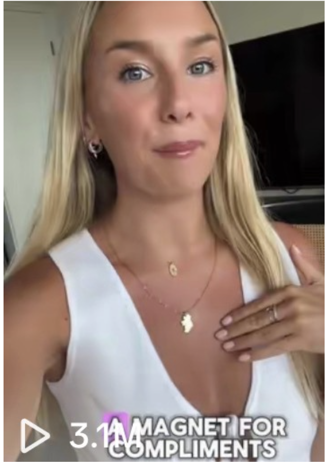 A woman with long blonde hair and blue eyes wearing a white sleeveless top, jewelry including earrings, a necklace, and a ring, posing for a selfie in front of a black television or monitor, with text overlay 'MAGNET FOR COMPLIMENTS' and a play button icon.