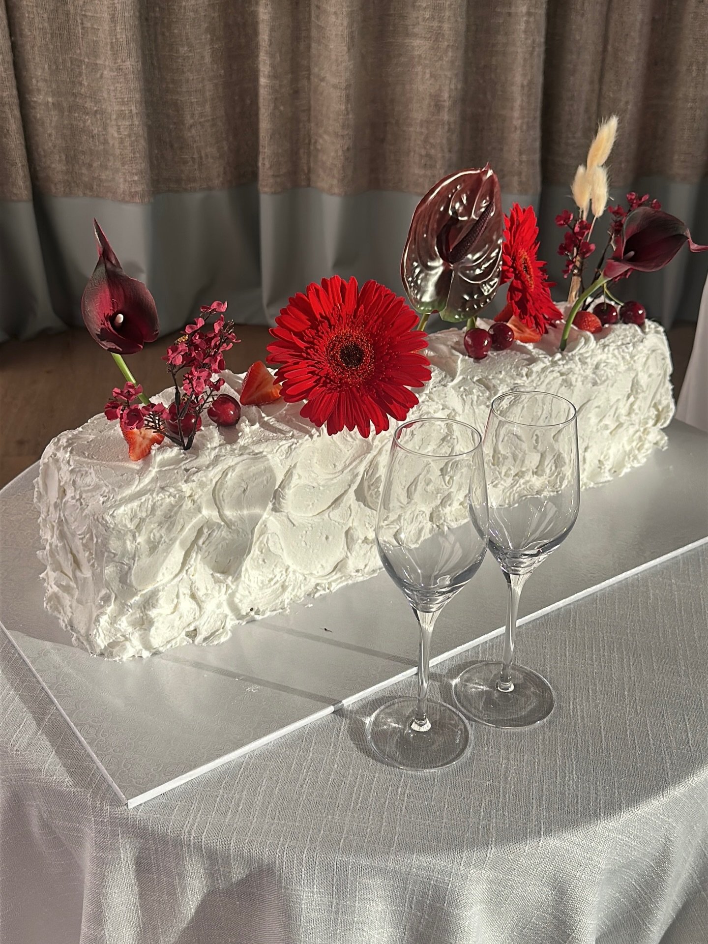 Elegant rectangular cake decorated with white frosting, red flowers, dark foliage, and Christmas-themed accents, displayed on a white stand with two empty wine glasses in front.