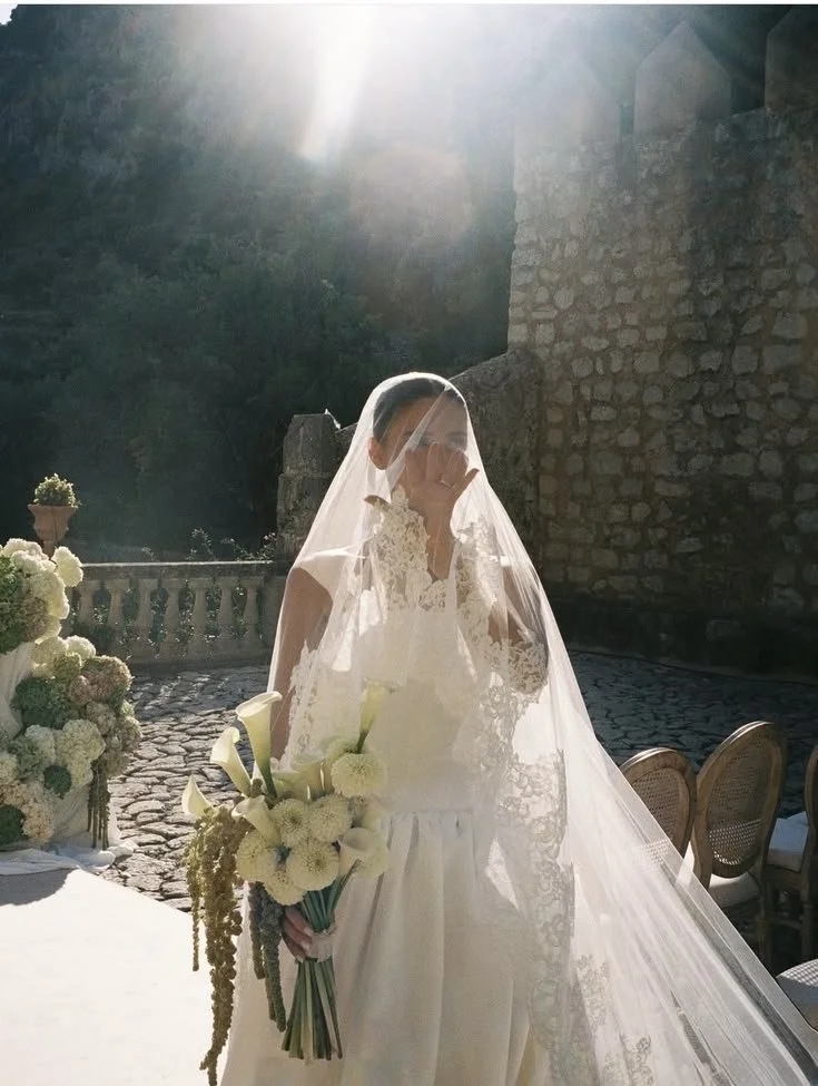 A bride in a white wedding dress and veil holding a bouquet of white flowers, standing outdoors in sunlight, with stone walls and patio furniture in the background.
