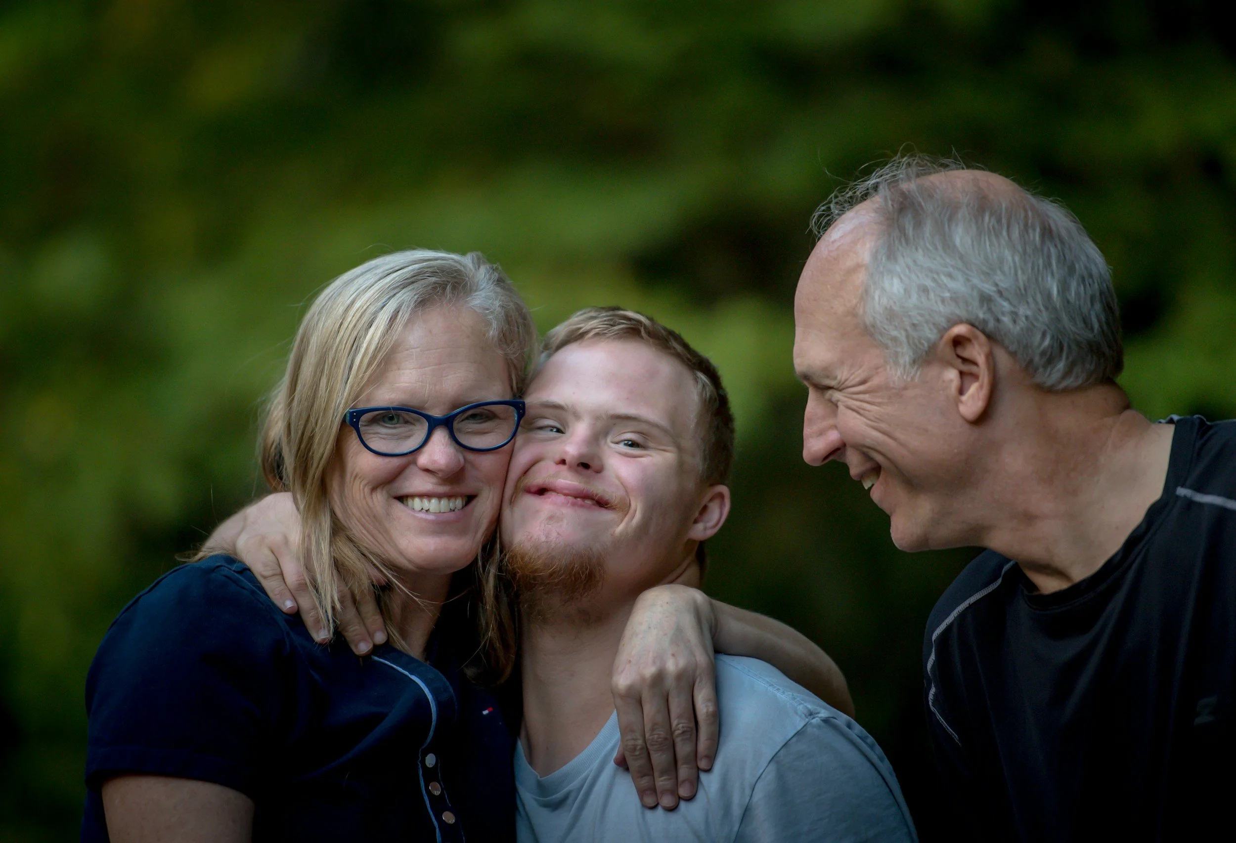 A happy family of three outdoors, smiling and hugging against a blurred green background.