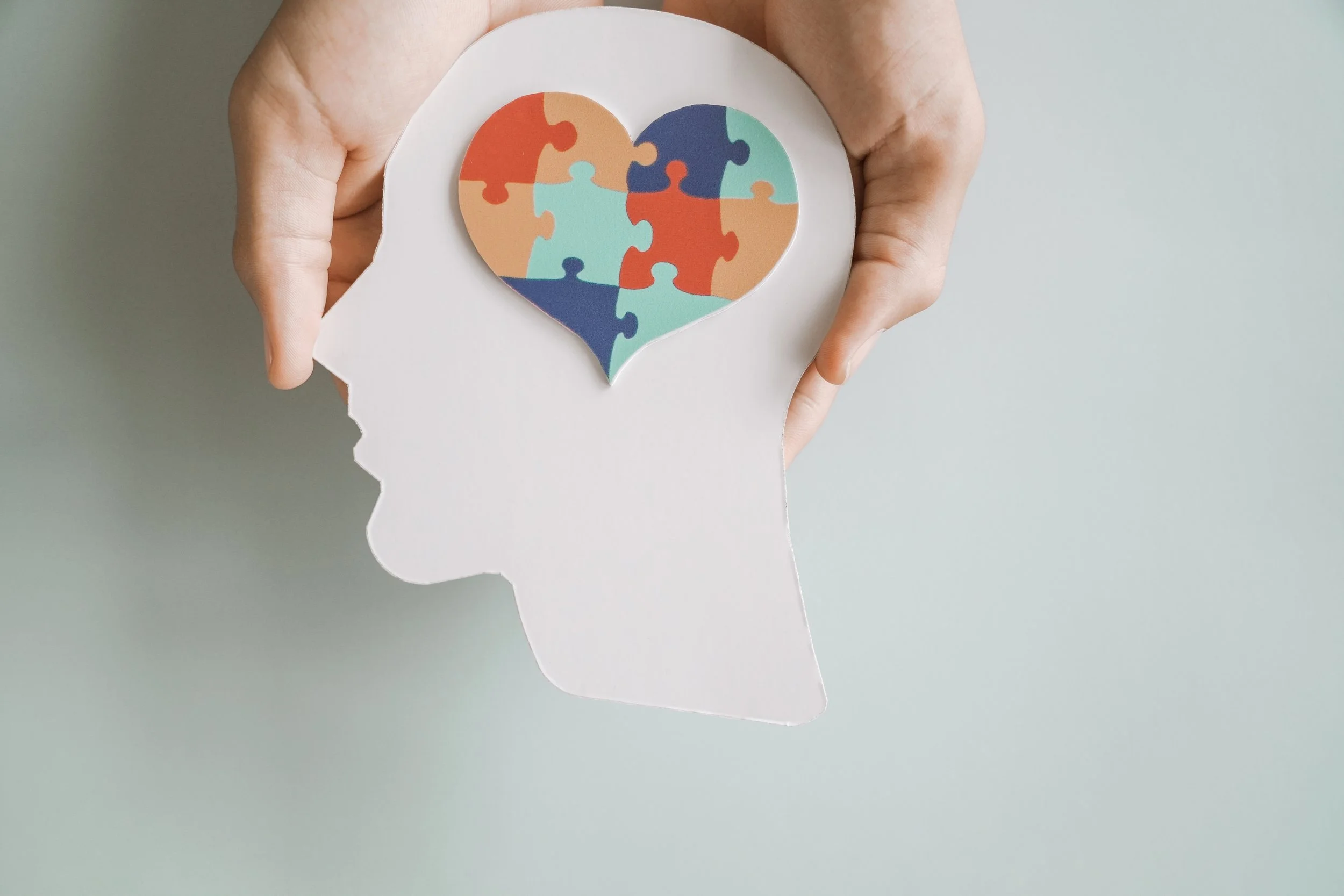 A white paper cutout of a human head silhouette holding a paper heart with puzzle pieces, placed over the head's brain area, symbolizing mental health or cognition.