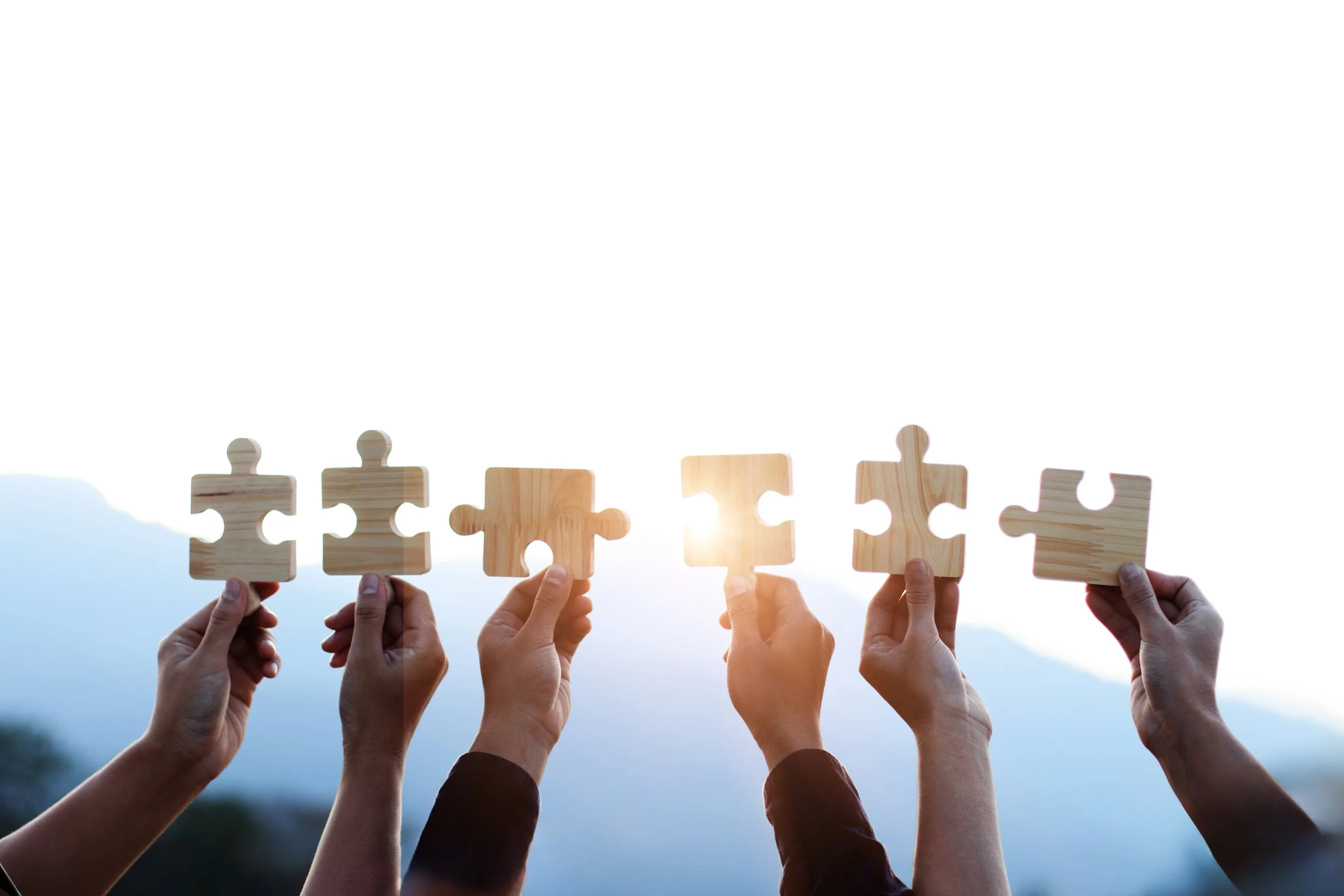 Six hands holding wooden puzzle pieces in the sky with mountain in the background, sunlight shining between the pieces, symbolizing teamwork or problem-solving.