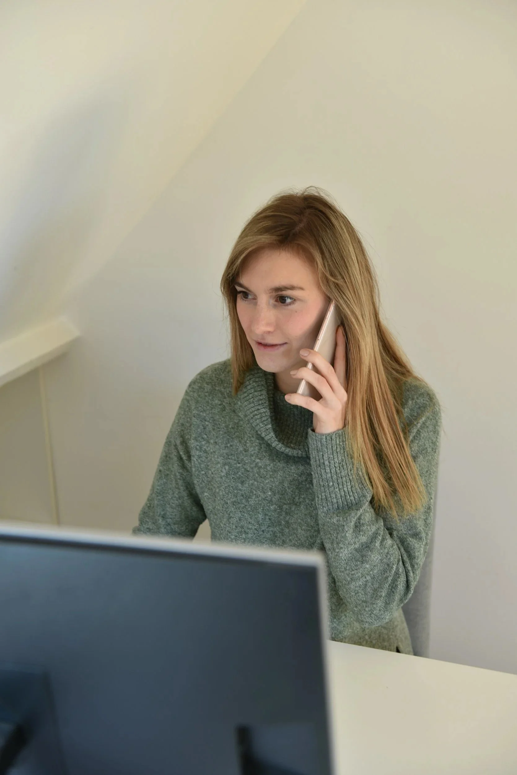 A woman with long red hair wearing a gray sweater talking on a phone while seated at a desk in front of a computer monitor.