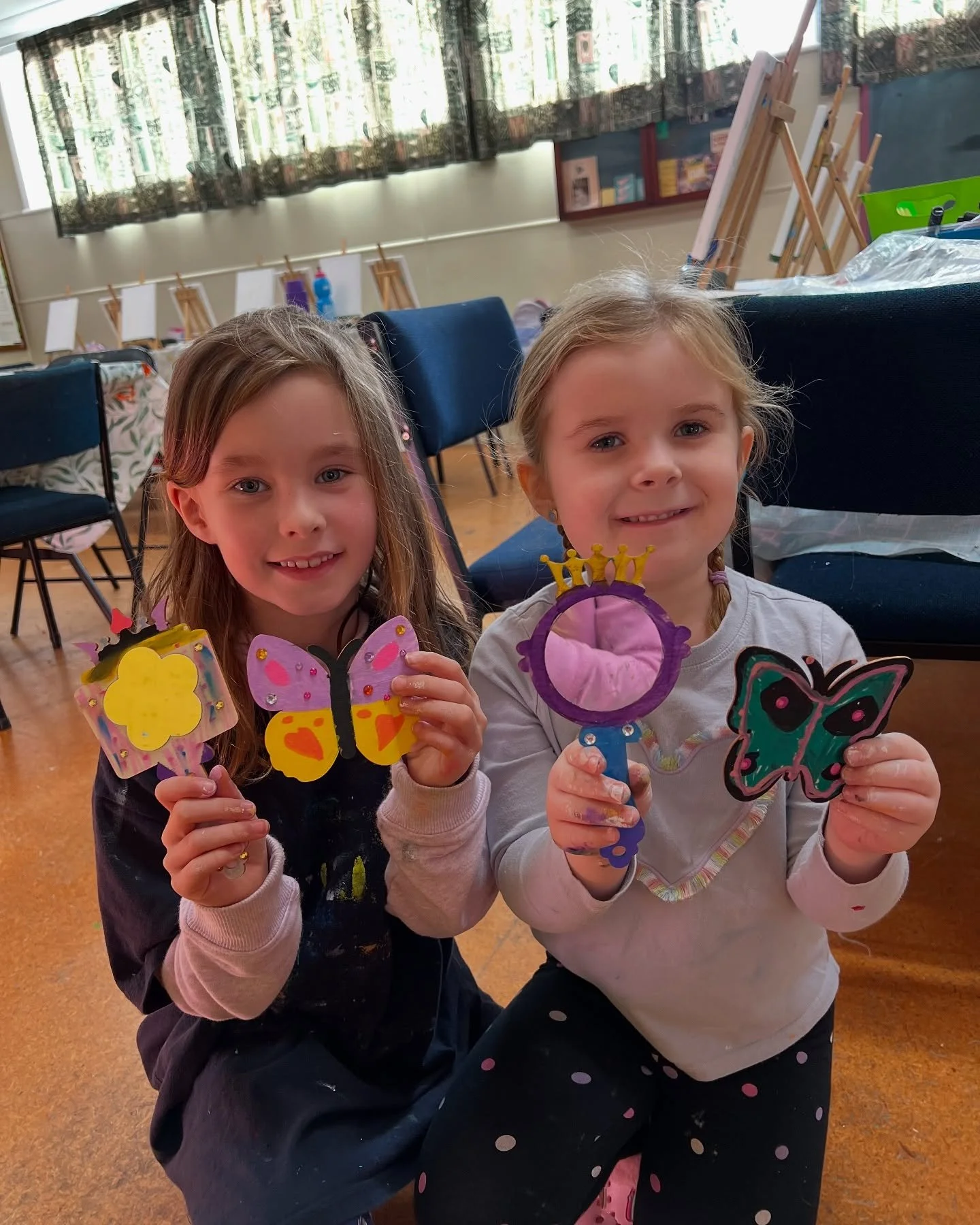 Two young girls smiling and holding colorful butterfly and crown crafts in a classroom.