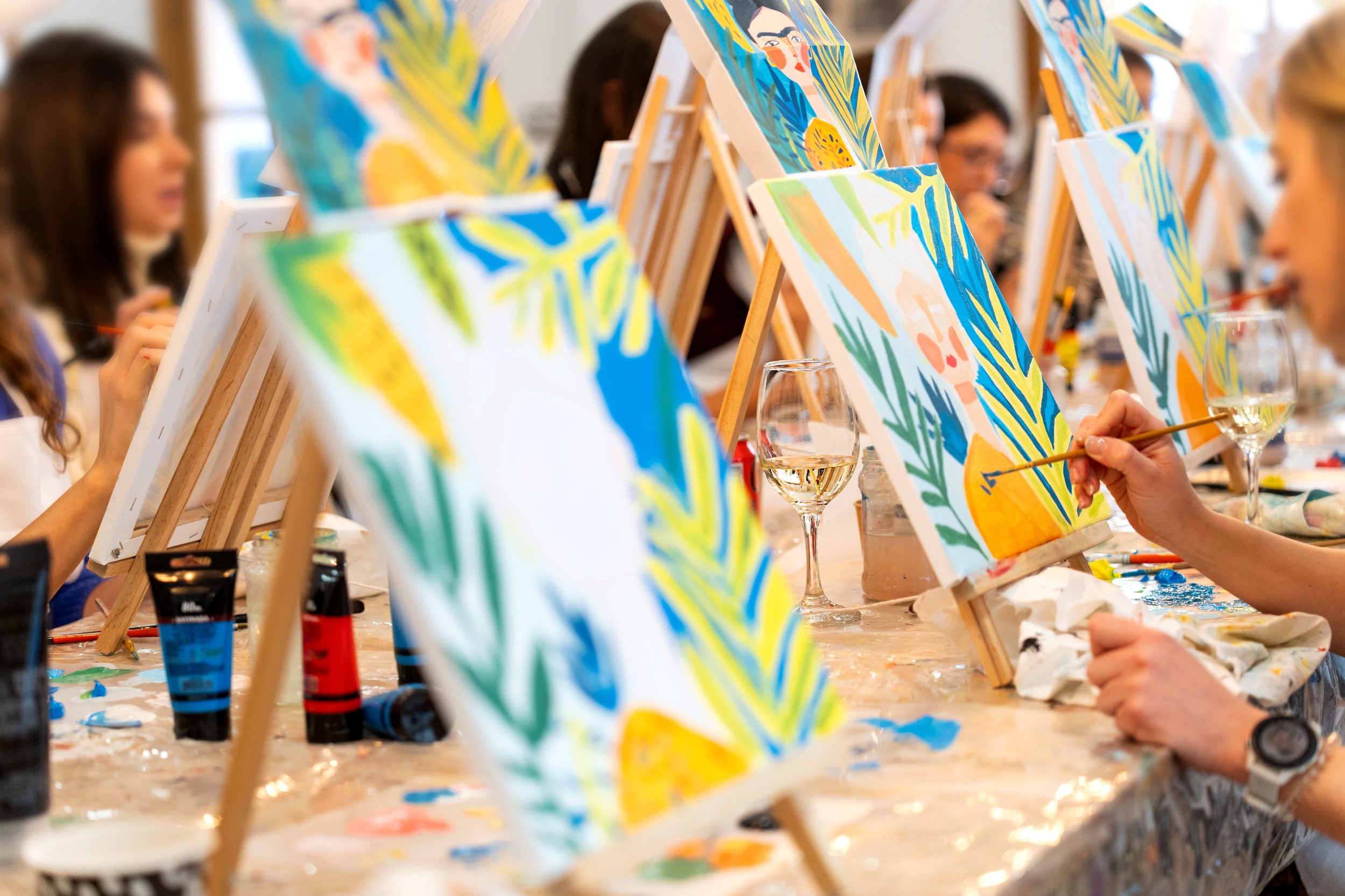 People participating in a group painting class, working on colorful landscapes on canvases at a long table in an art studio.