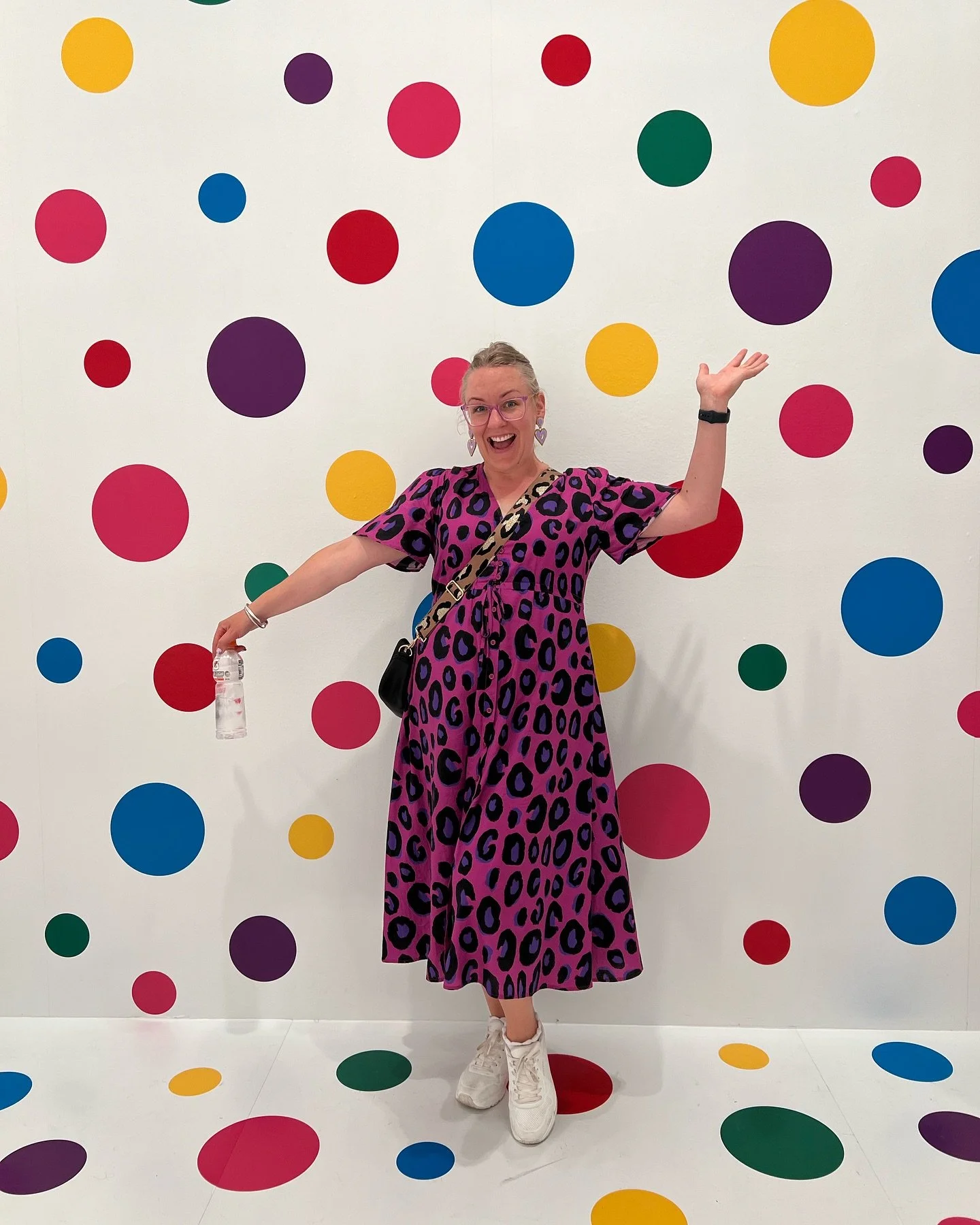 A woman in a pink and purple leopard print dress with white sneakers, glasses, and earrings standing in front of a white wall with colorful polka dots, smiling, holding a water bottle, with her arms raised.