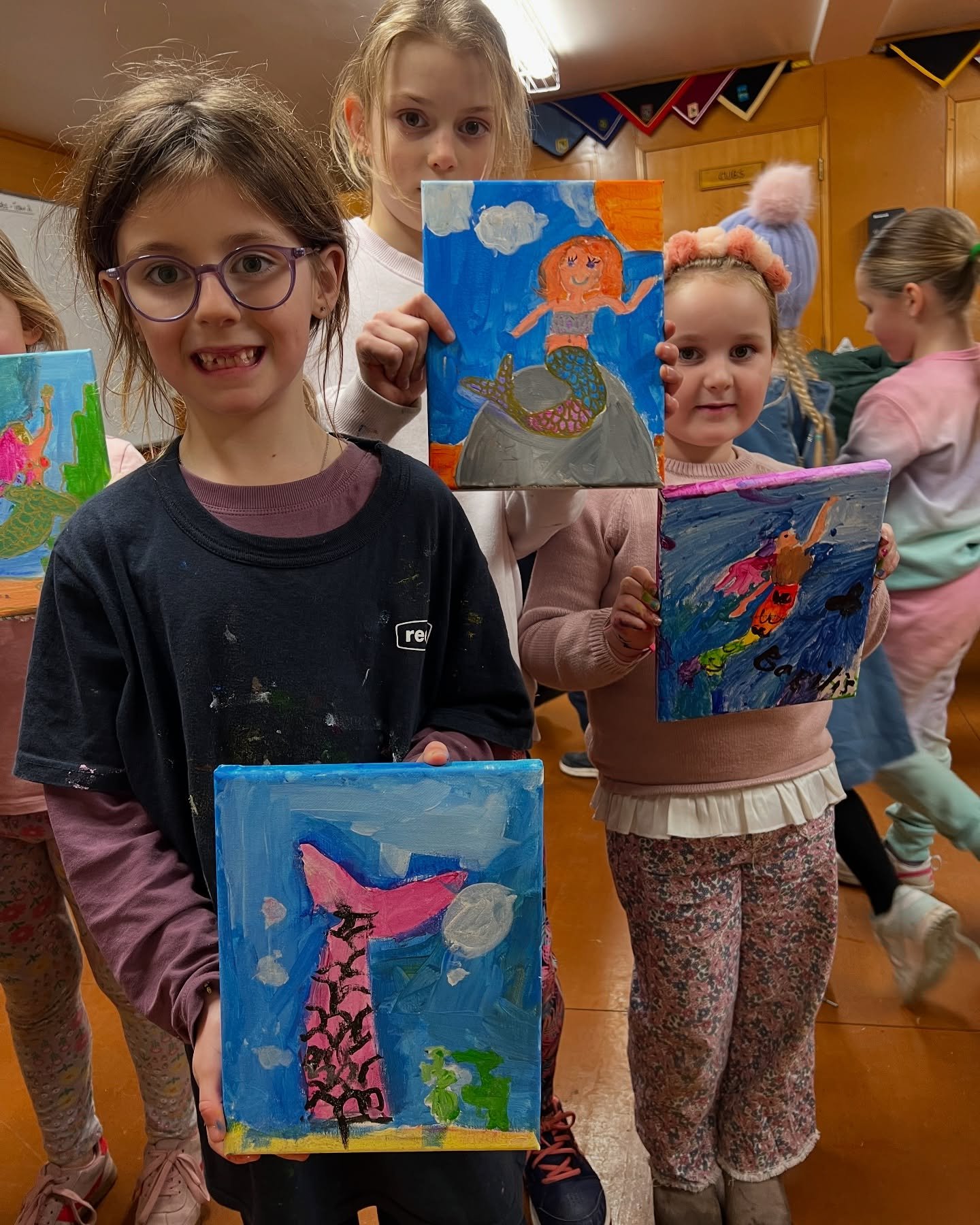 Four young girls displaying colorful paintings of mermaids, some holding their artwork, in a classroom setting.