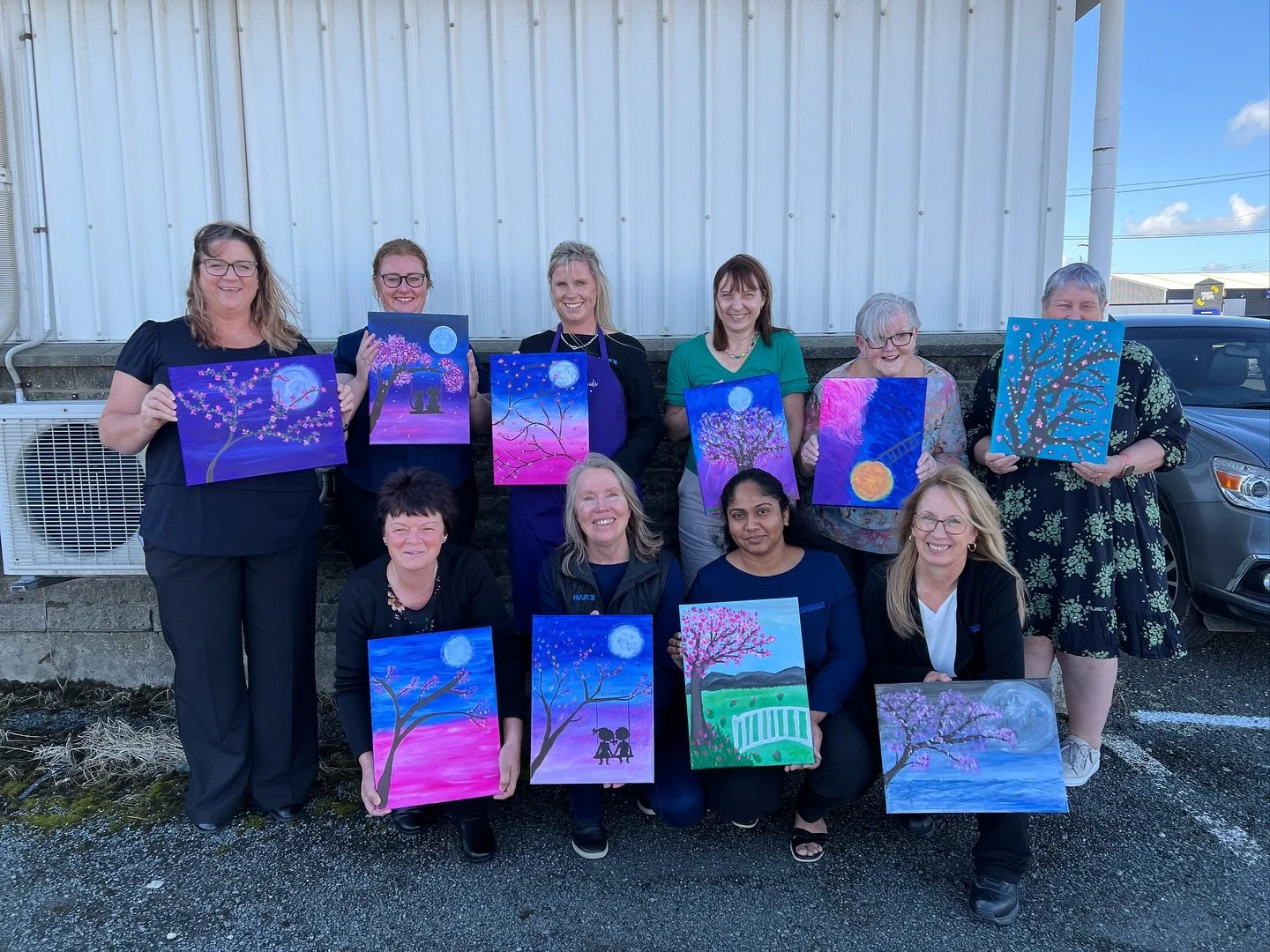 Group of women holding paintings with moon and cherry blossom themes outdoors.