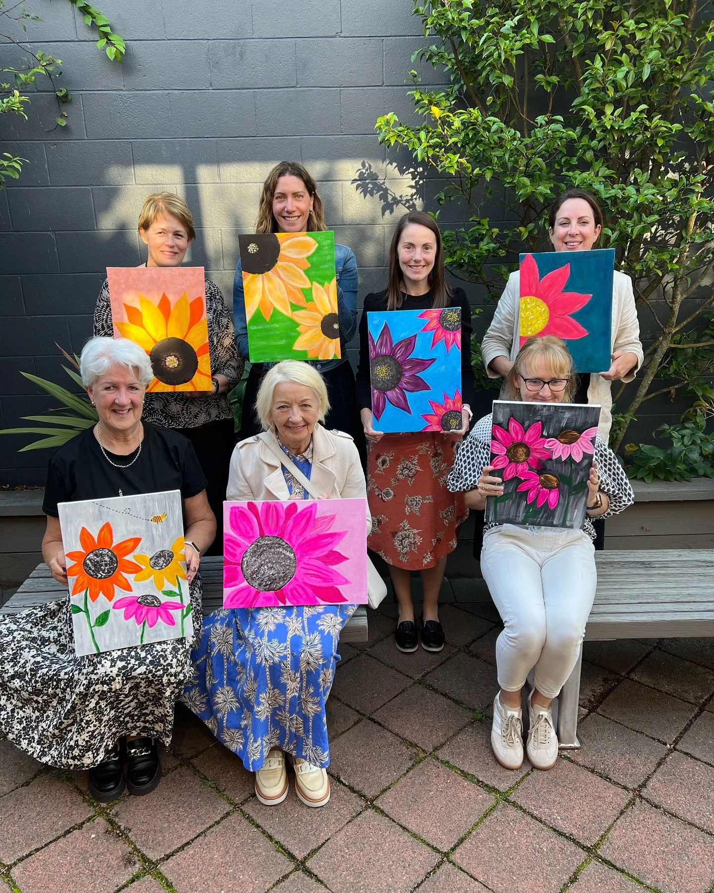 A group of nine women posing outdoors, each holding colorful paintings of large flowers. They are sitting and standing in front of a dark wall with green plants and trees.