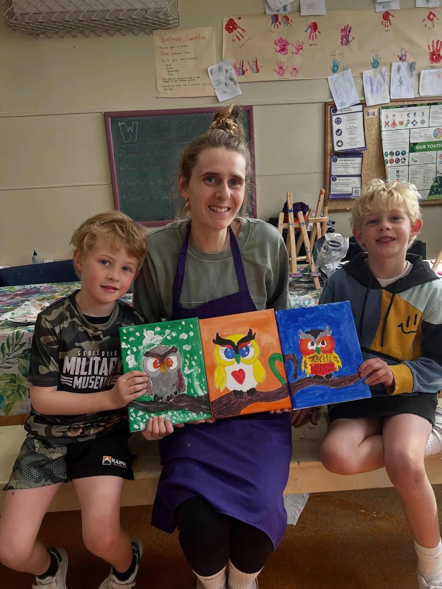 A woman and two young boys sitting on a bench in an art classroom, holding three colorful owl paintings they made. The woman is wearing a purple apron and has her hair up in a bun, smiling at the camera. The boys are also smiling, one wearing a camouflage shirt and the other in a hoodie, both showing their paintings of owls with different backgrounds.