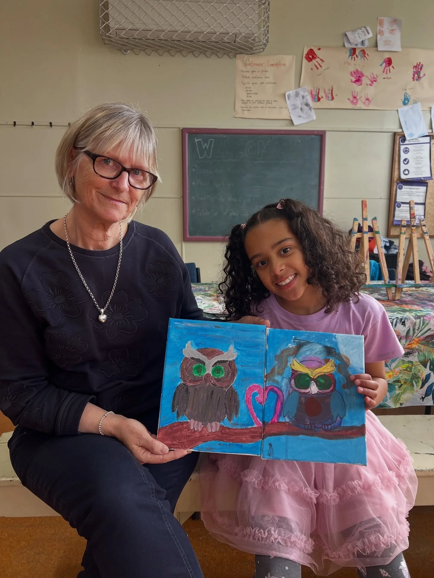 A woman and a girl sitting together in a classroom, holding up drawn pictures of owls. The woman has short blond hair, glasses, and is wearing a black shirt with floral embroidery. The girl has curly hair, is smiling, and is wearing a pink dress.