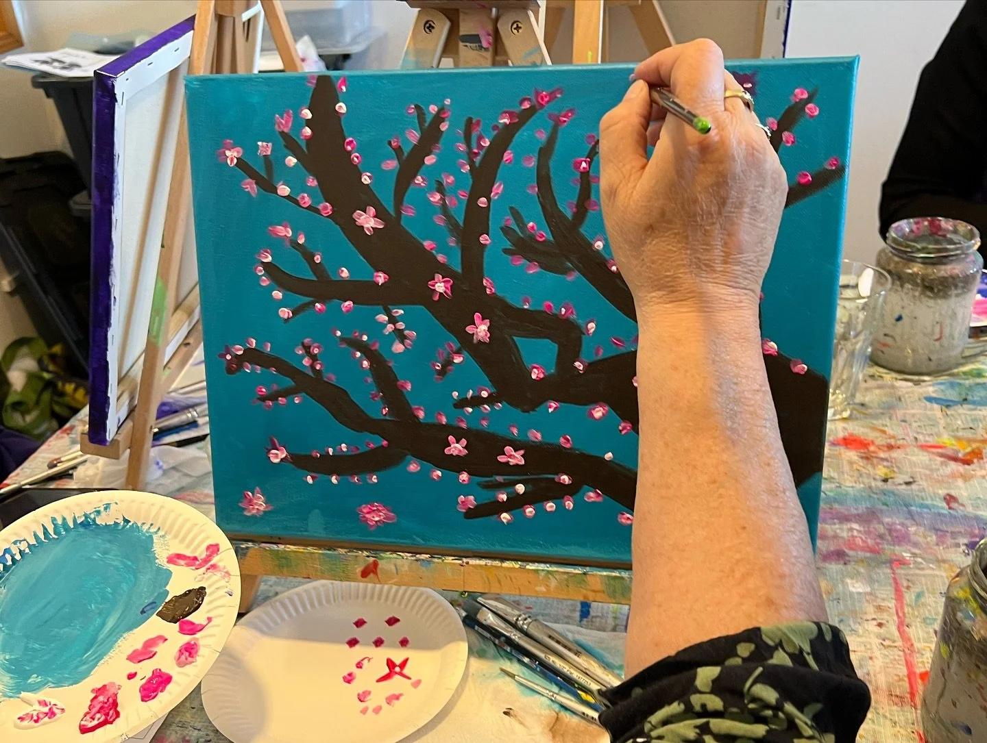 Person painting a cherry blossom tree on a canvas, with pink flowers and a blue background, using small brushes and pink and white paint.