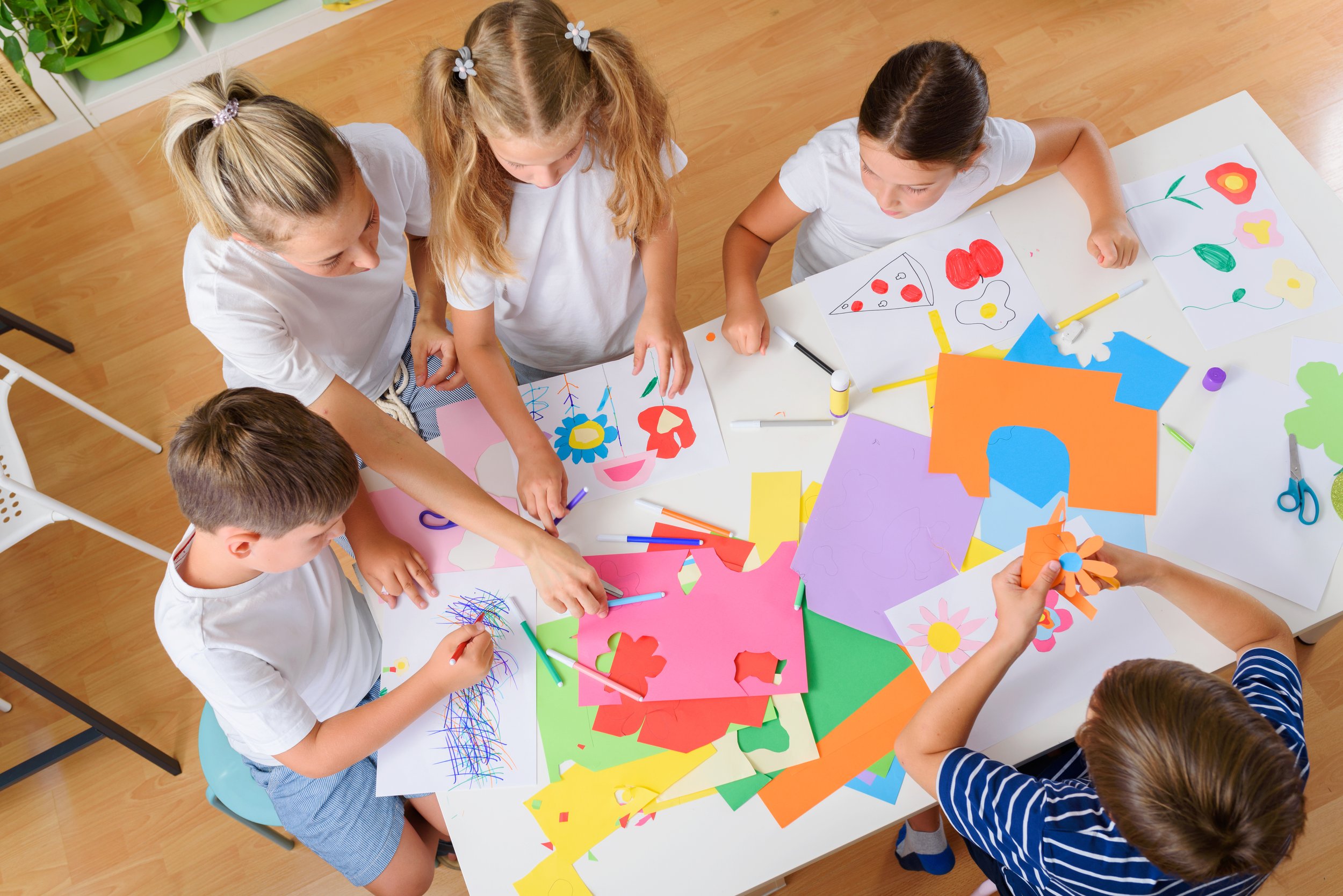 Children engaging in arts and crafts, drawing and cutting colored paper with scissors at a table.