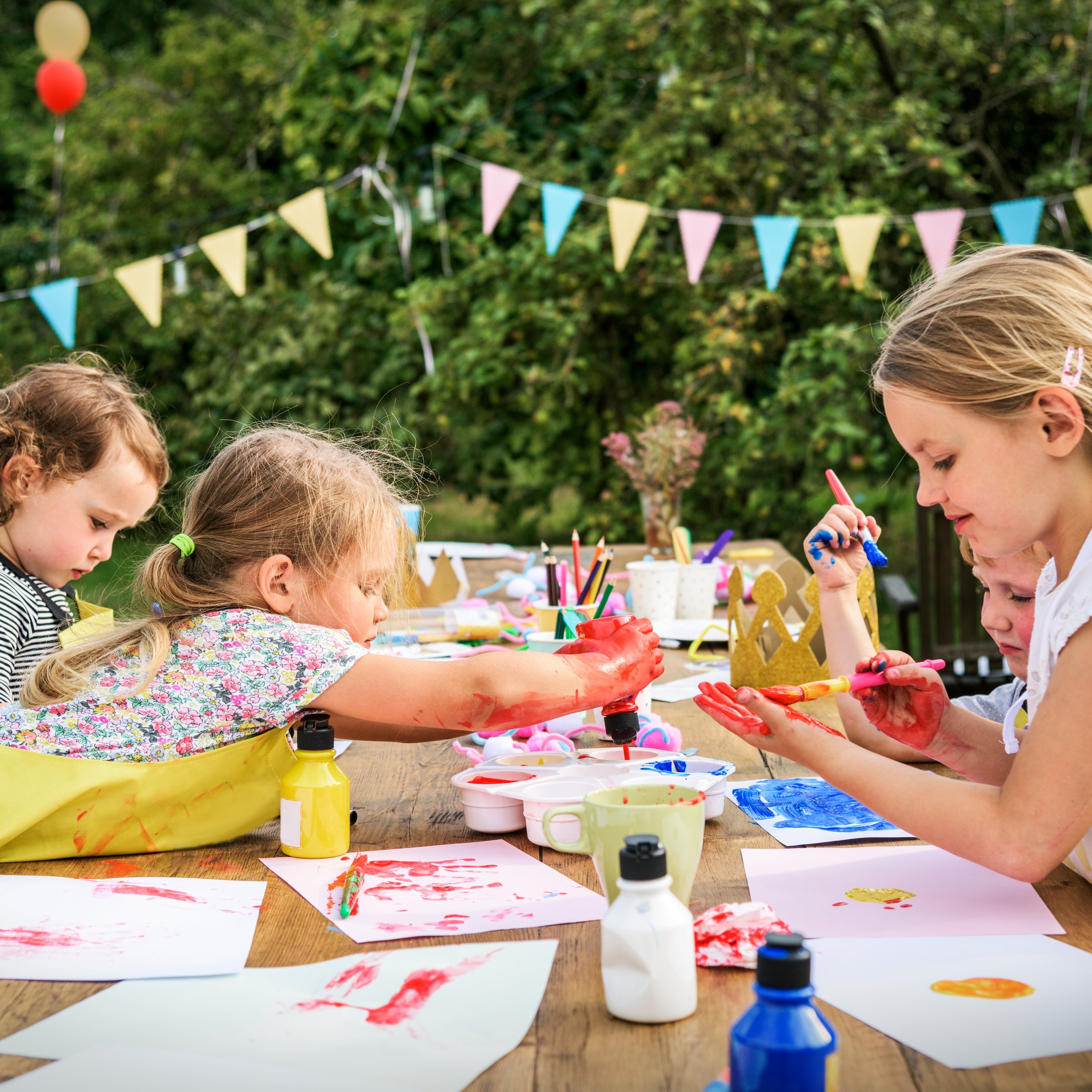 Children painting and drawing at an outdoor table decorated with colorful flags, with art supplies and artwork on the table.