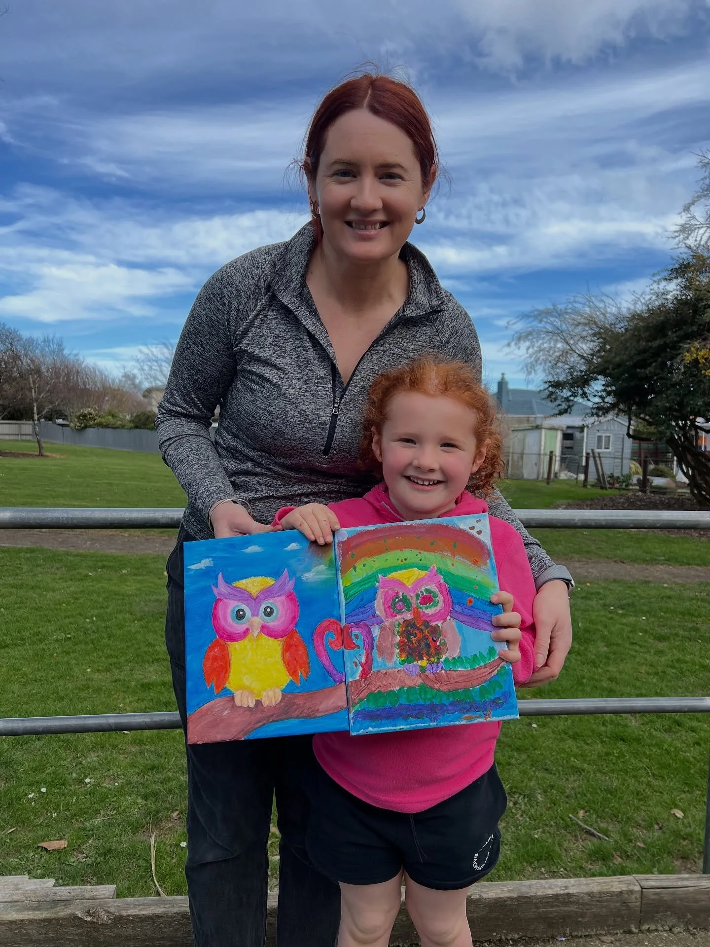 A woman and a girl holding colorful owl paintings outdoors on a grassy area with trees and houses in the background.