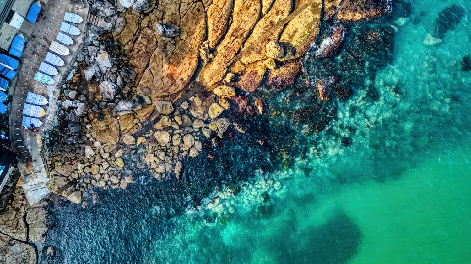 Aerial view of a rocky shoreline with turquoise water and a row of blue and white lounge chairs along the coast.