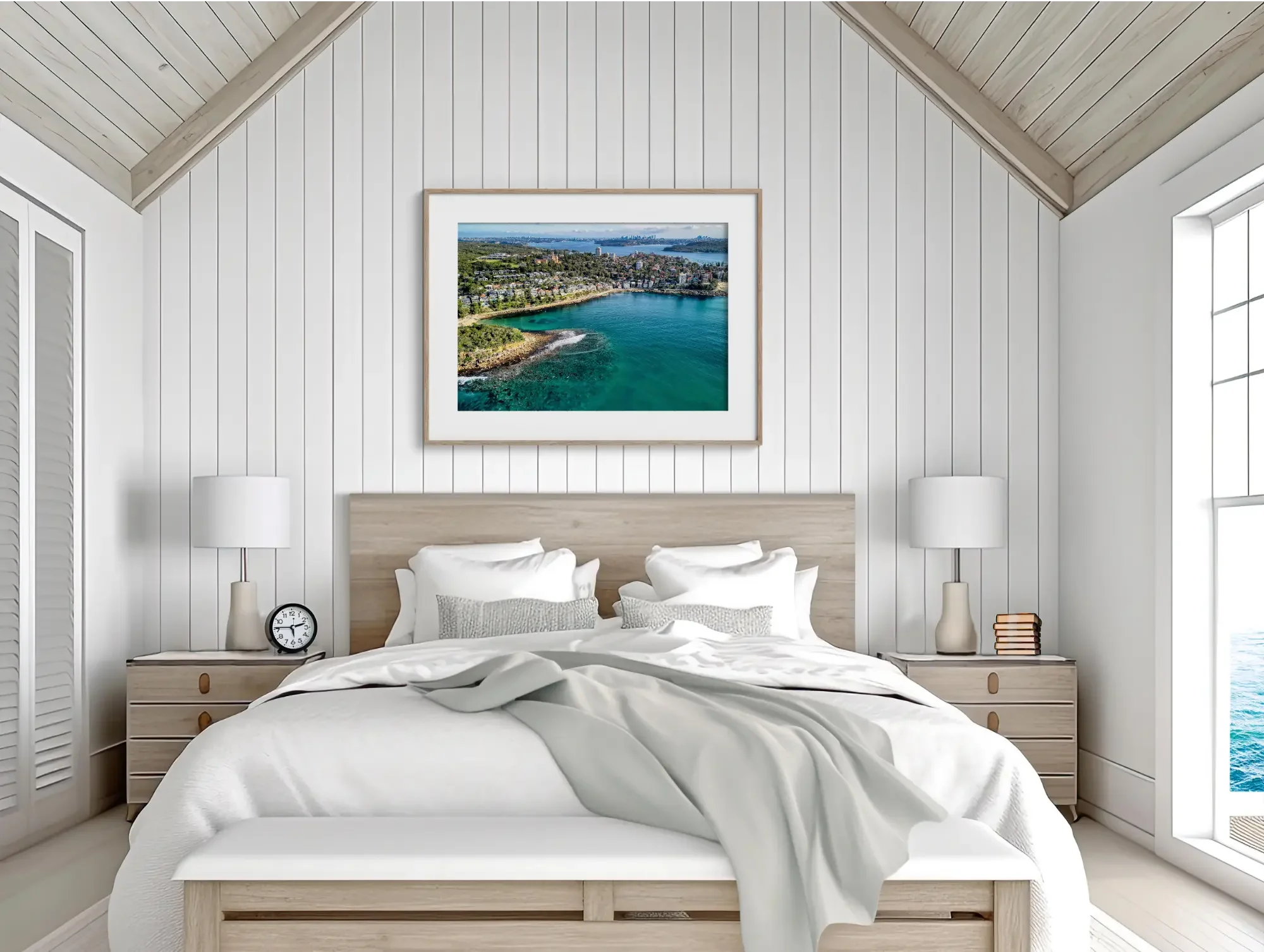 A cozy bedroom with white paneled walls and a slanted wooden ceiling. A framed aerial photo of a coastal city hangs above the bed. The bed has white linens, and there are white lamps on bedside tables.