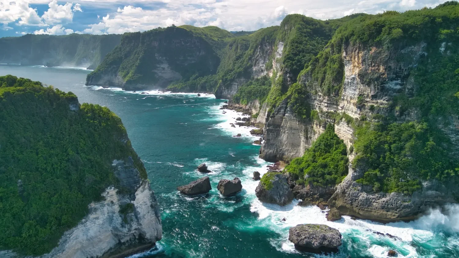 Coastal cliffs covered with green vegetation overlooking the ocean with rocks and waves crashing against the shoreline on a partly cloudy day.