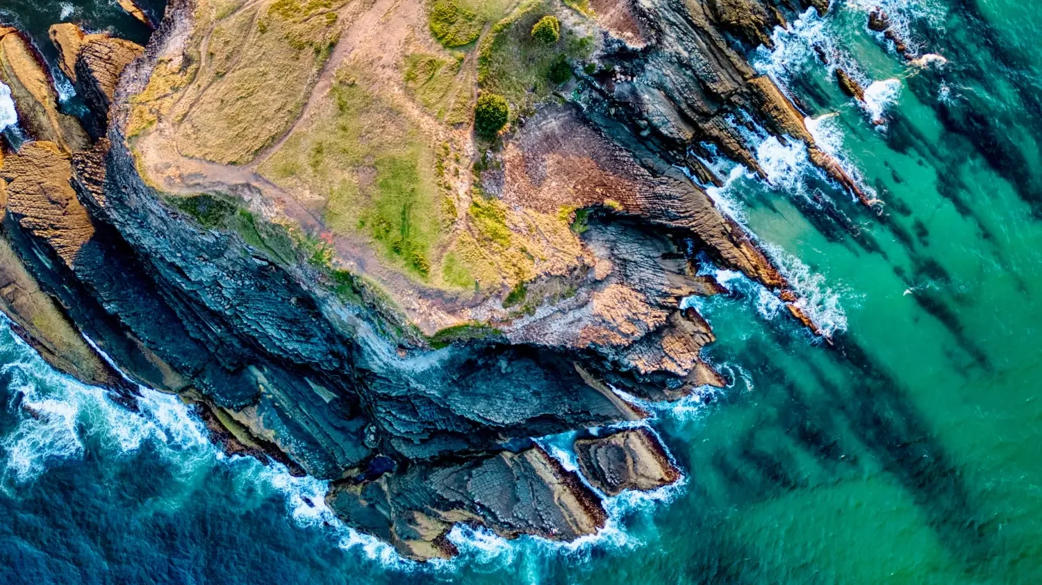 Aerial view of rocky coastline with cliffs, green grassy patches, and ocean waves crashing against the rocks.