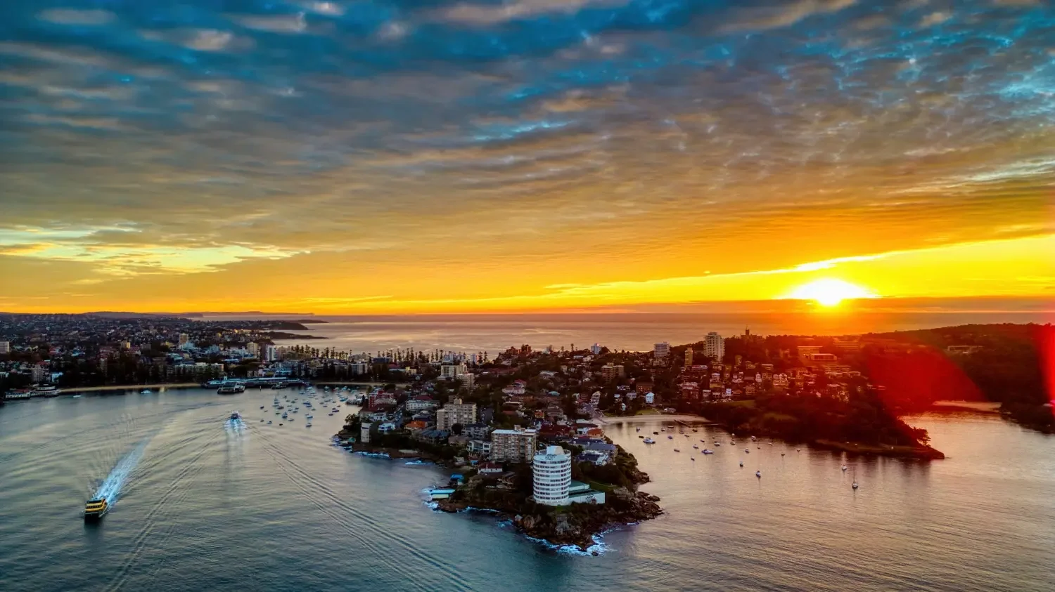 Aerial view of a coastal city at sunset, featuring a harbor with boats, a small island with buildings, and a city skyline with various high-rise and low-rise buildings under a vibrant sky with orange, yellow, and blue hues.