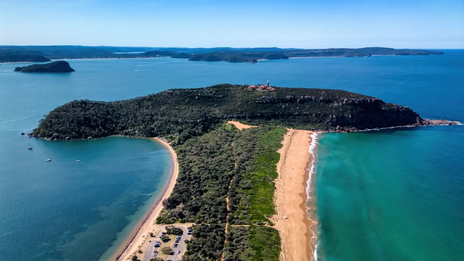 Aerial view of a lush, green island with a lighthouse on top, surrounded by blue ocean waters with beaches along the coast, and distant islands in the background.