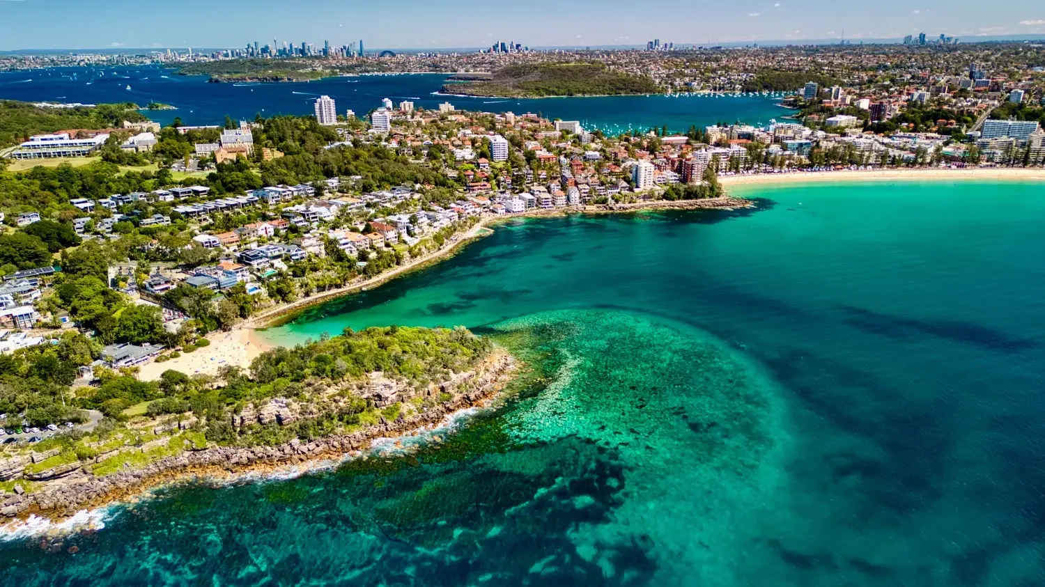 Aerial view of a coastal city with turquoise waters, sandy beaches, residential area with trees, and a city skyline in the distance.
