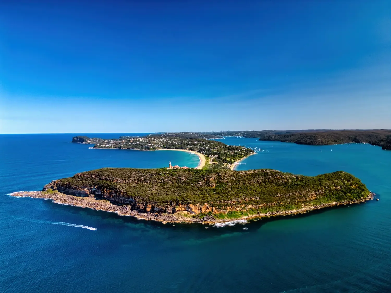 Aerial view of a coastal island with rocky cliffs, green vegetation, and a lighthouse near the shoreline, surrounded by blue ocean water with a boat nearby and a distant shoreline with residential area and boats.