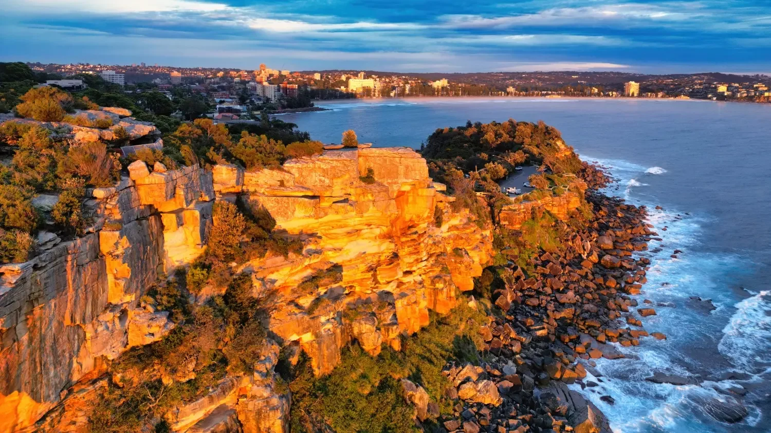 View of rocky coastal cliffs illuminated by sunlight near the ocean with a cityscape in the distance under a partly cloudy sky.