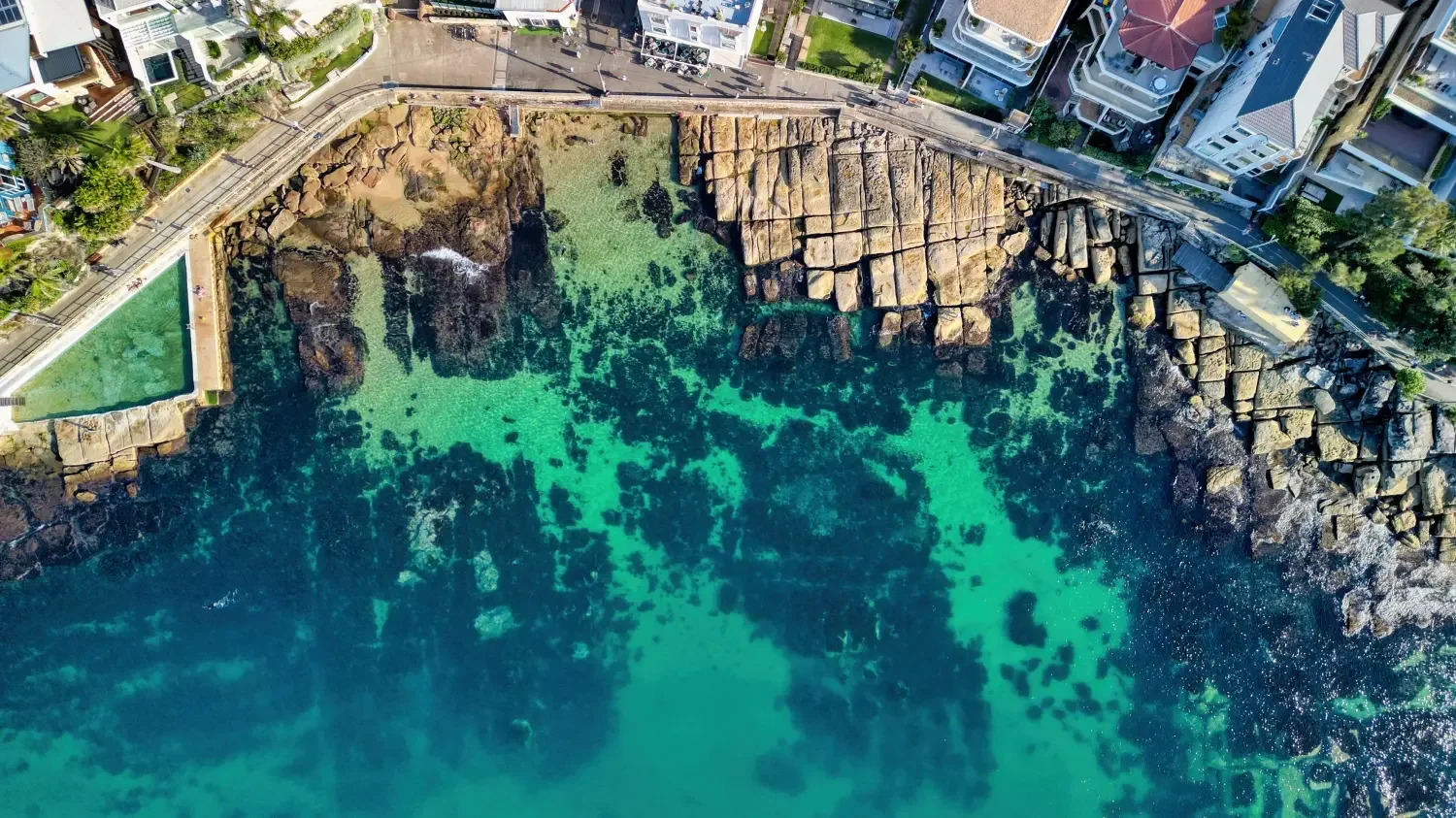 Aerial view of a rocky shoreline with turquoise water, bordered by urban buildings and a road with a sidewalk.