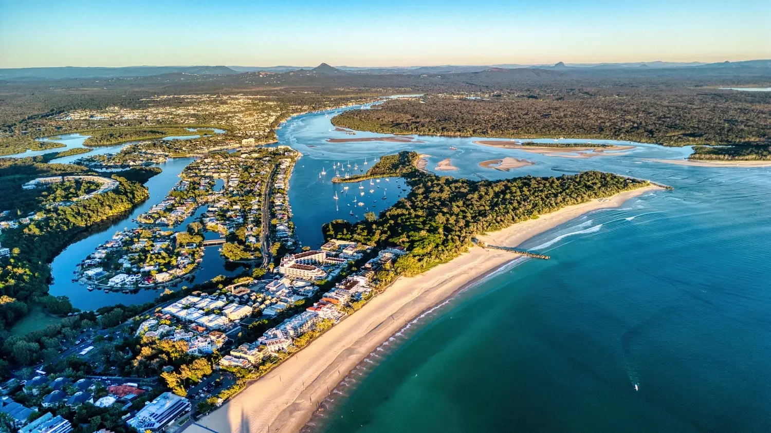 Aerial view of a coastal area showing a beach, a river, residential and commercial buildings, boats on the water, and green forested areas with distant hills.