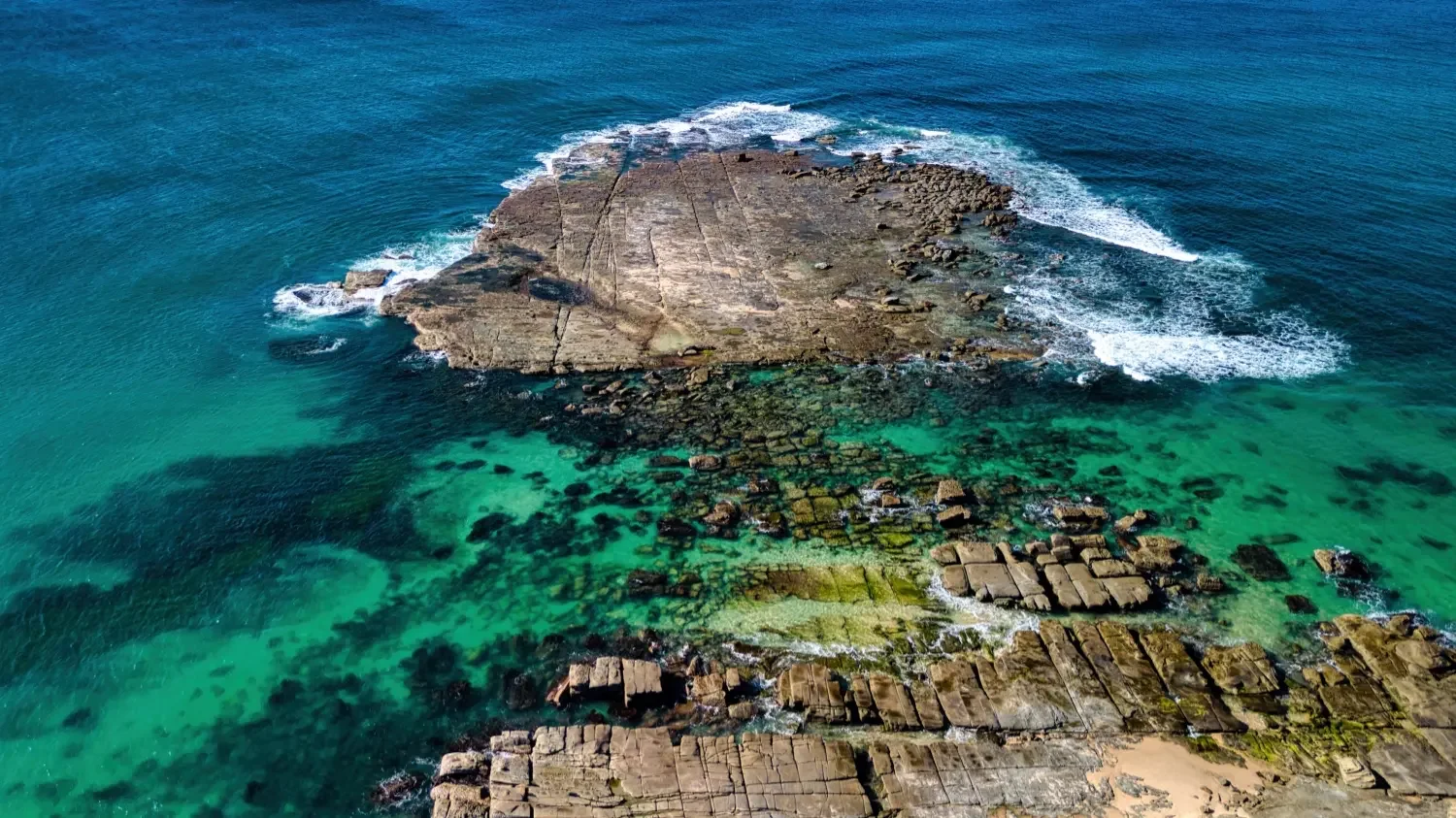 Aerial view of a rocky coastline with clear turquoise water, waves crashing against the rocks, and algae-covered stones along the shore.