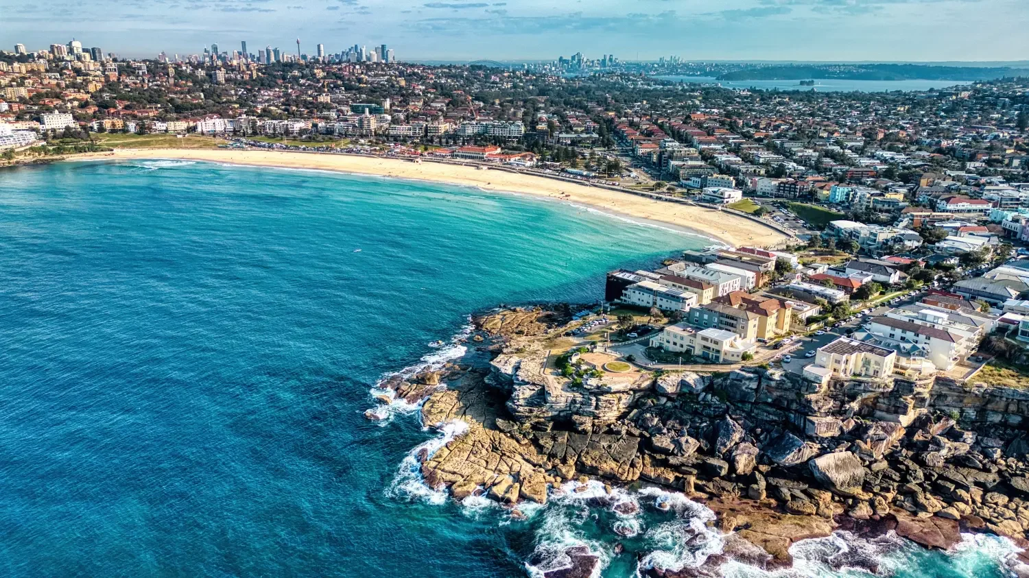 Aerial view of a city beach with turquoise water, sandy shoreline, and rocky coastline, with residential buildings and city skyline in the background.