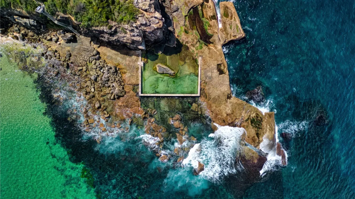 Aerial view of a rocky coastline with waves crashing against rocks, a small pool on the rocks, and greenery on the land.