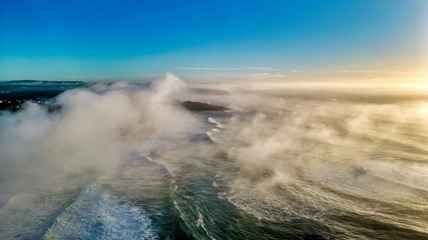 Aerial view of a large ocean wave crashing with spray and mist, with land and a cloudy sky in the background at sunrise or sunset.
