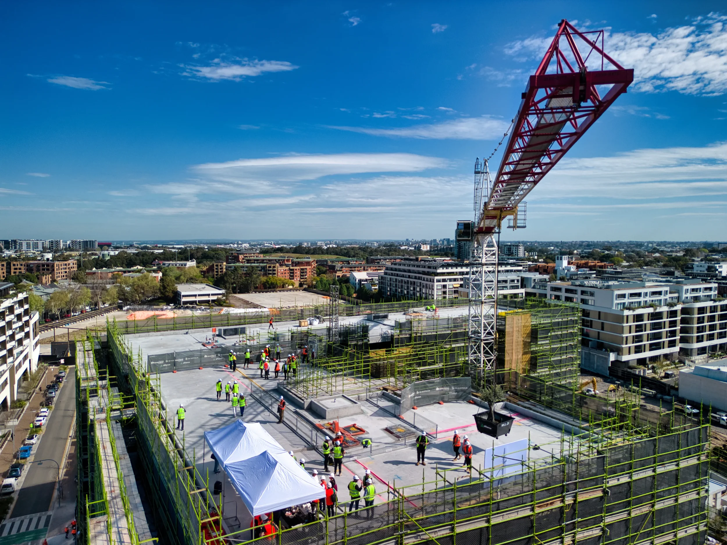 Construction site on a high-rise building with workers wearing safety vests and helmets, a crane, and tents set up, cityscape in the background under a partly cloudy sky.