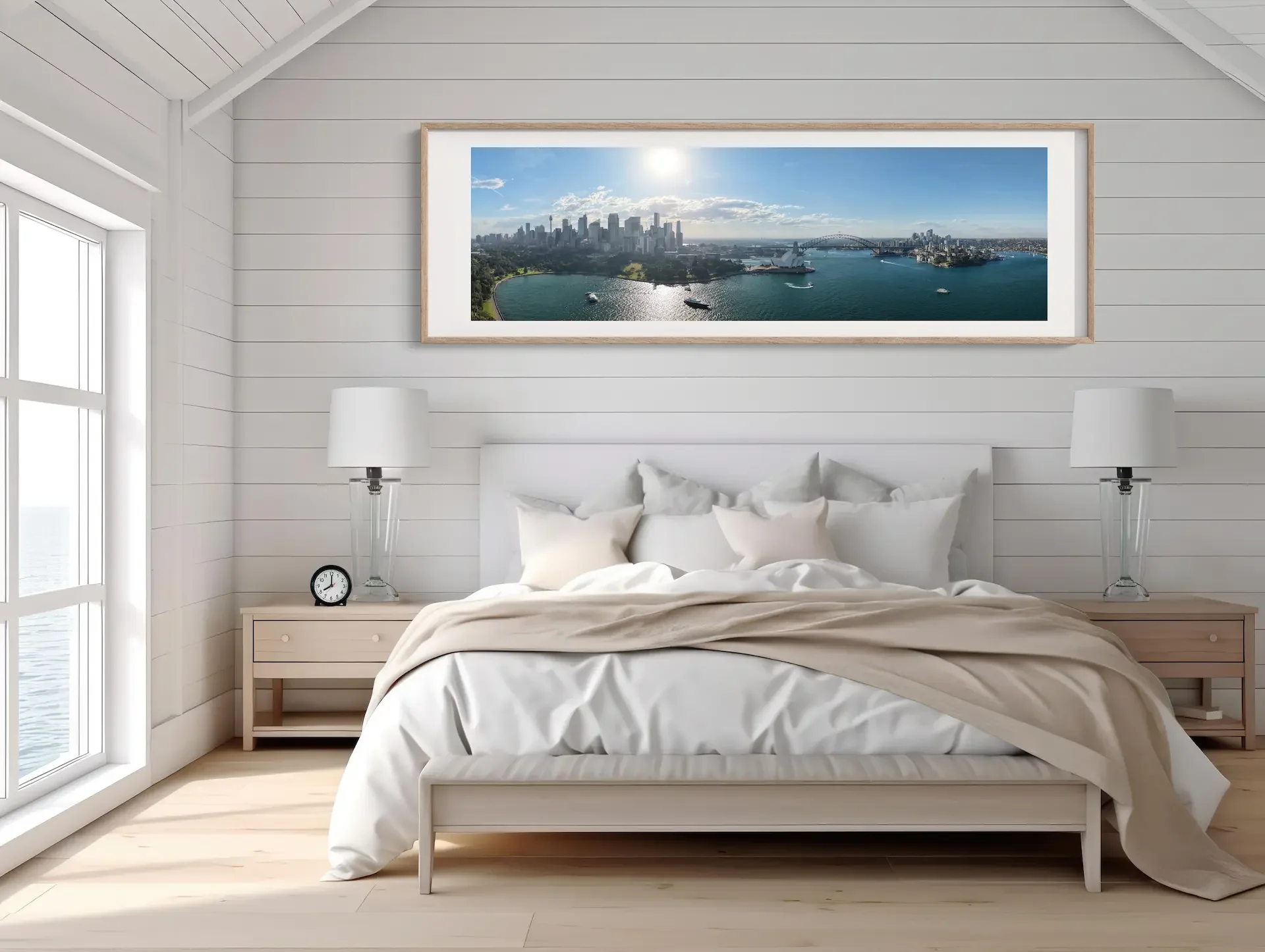 A bedroom with white walls and a white bed with white bedding. There are two nightstands with lamps, a clock, and a large framed photograph of a city skyline and river view above the bed. Natural light is coming through a large window on the left.
