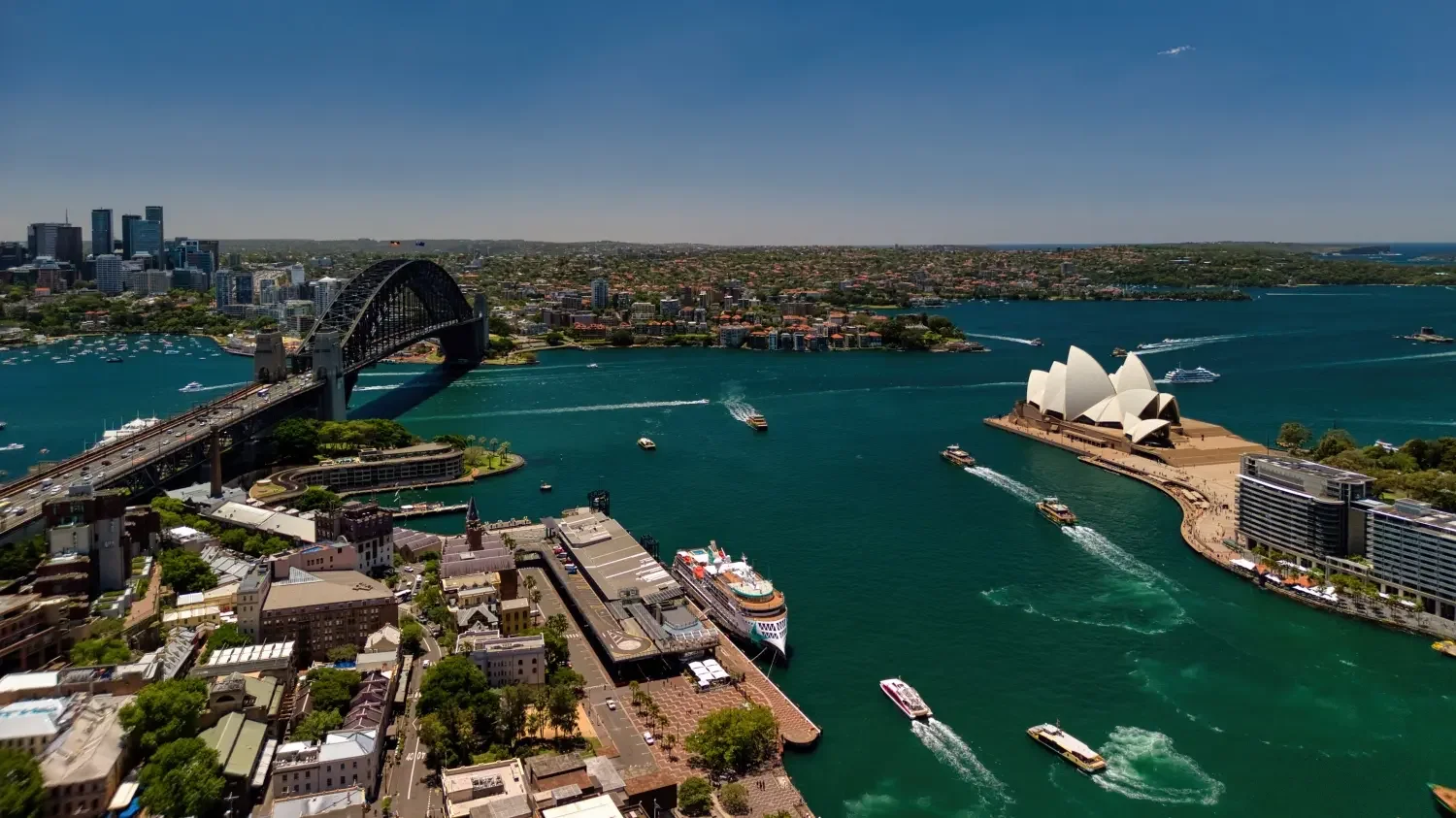 An aerial view of Sydney Harbour showcasing the Sydney Opera House, Harbour Bridge, boats on the water, and the city skyline in the background.