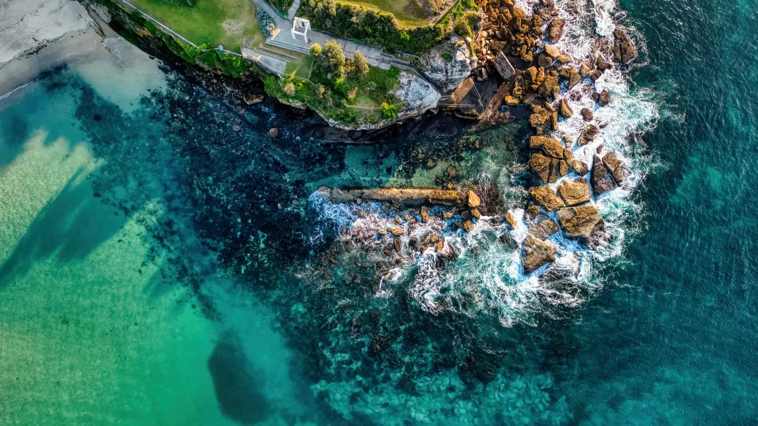 Aerial view of a coastal area showing rocks, seawater, and green grass with trees and a small concrete structure.