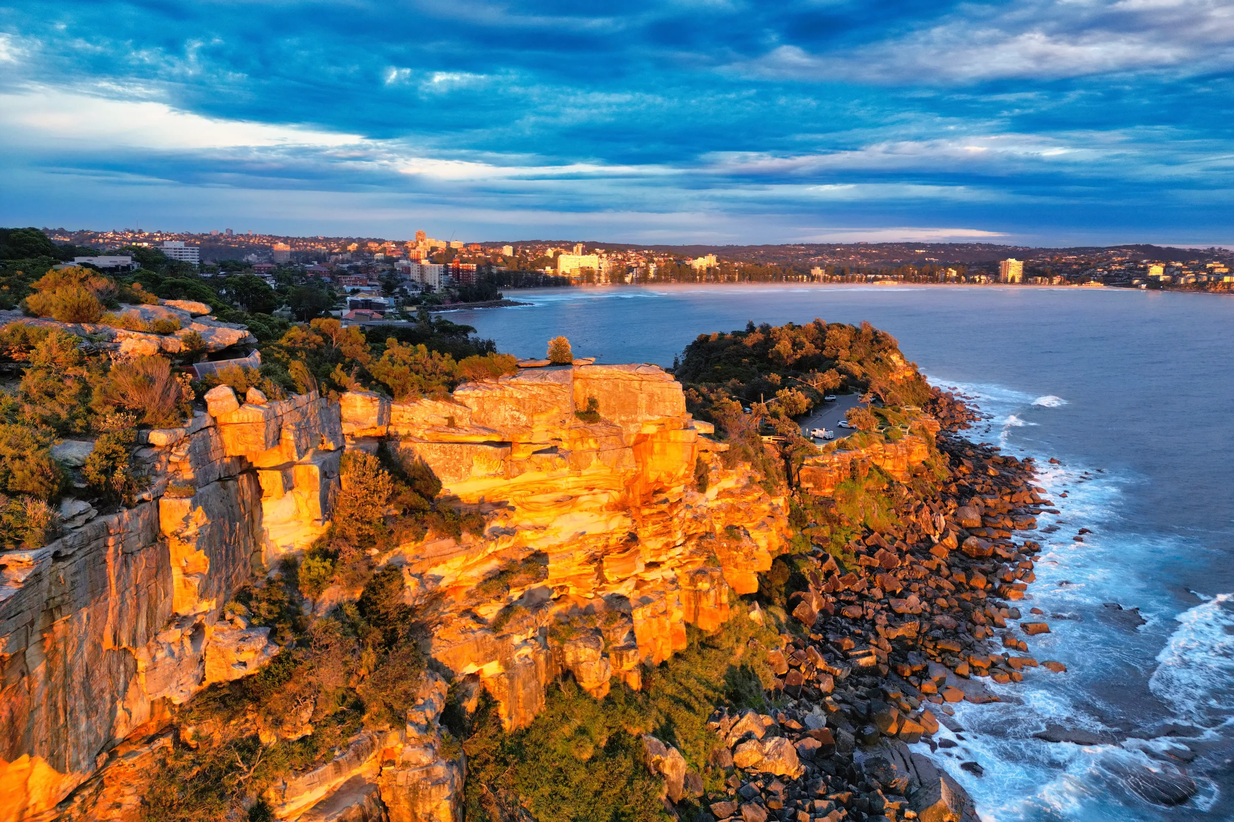 A coastal cityscape at sunset with rocky cliffs, greenery, and buildings along the shoreline under a partly cloudy sky.