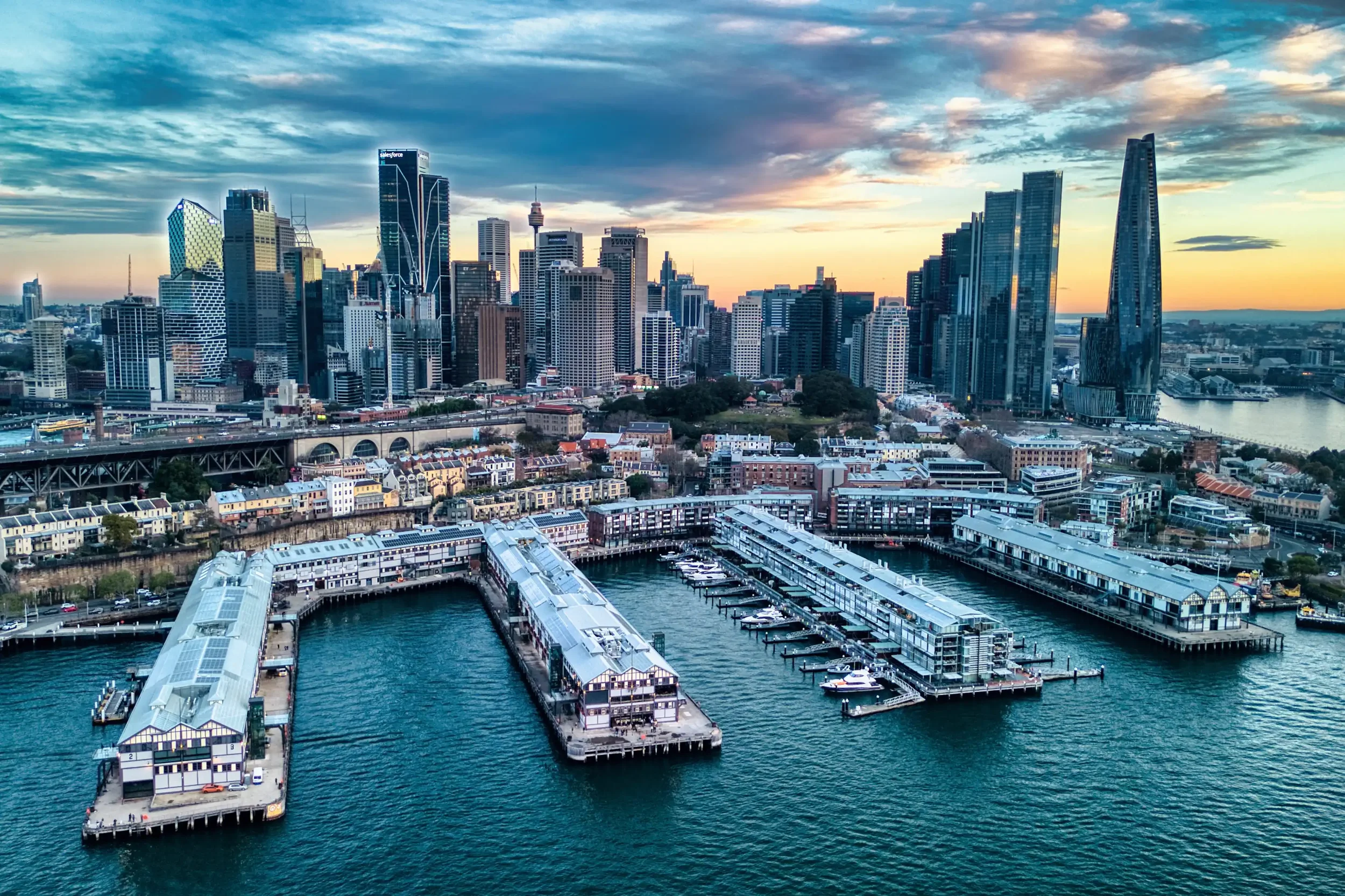 A city skyline at sunset featuring high-rise buildings, a waterfront with boats and a marina, with a partly cloudy sky overhead.